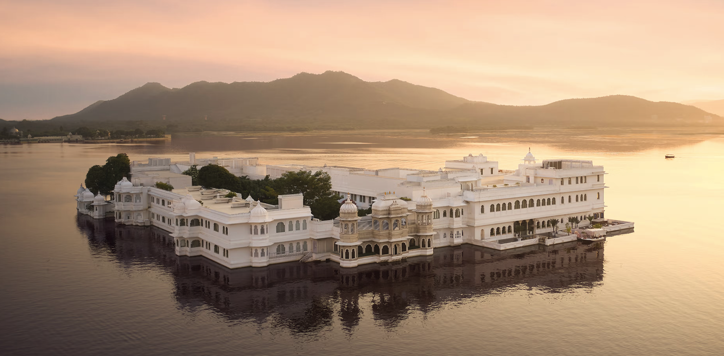 A white palace with traditional Indian architecture on a lake, reflecting in the water, with hills and a sunset sky in the background.