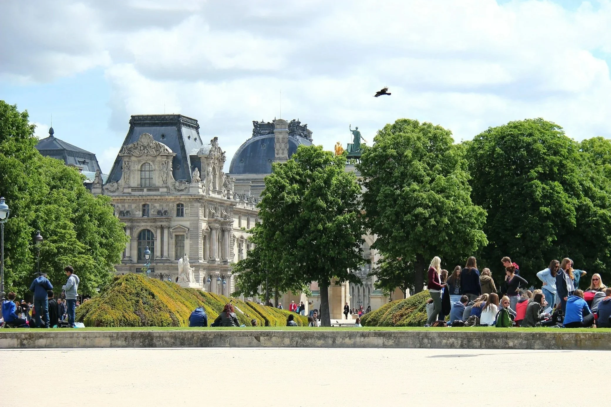 People sitting and standing in a park with green trees, in front of historic European-style buildings with ornate architecture, and a cloudy sky.
