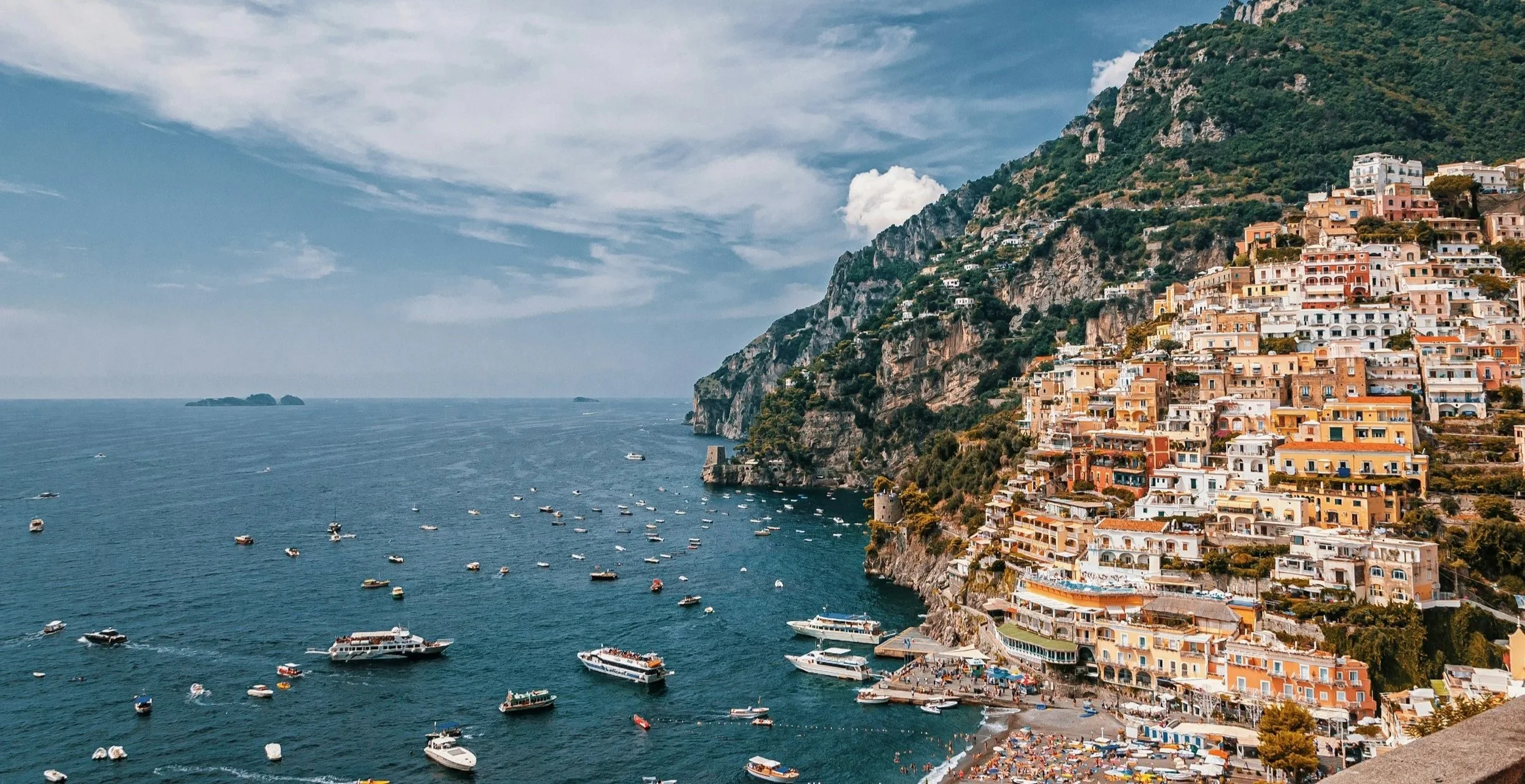 Colorful buildings on a steep hillside overlooking the Mediterranean Sea with boats docked in the water and a small beach below.