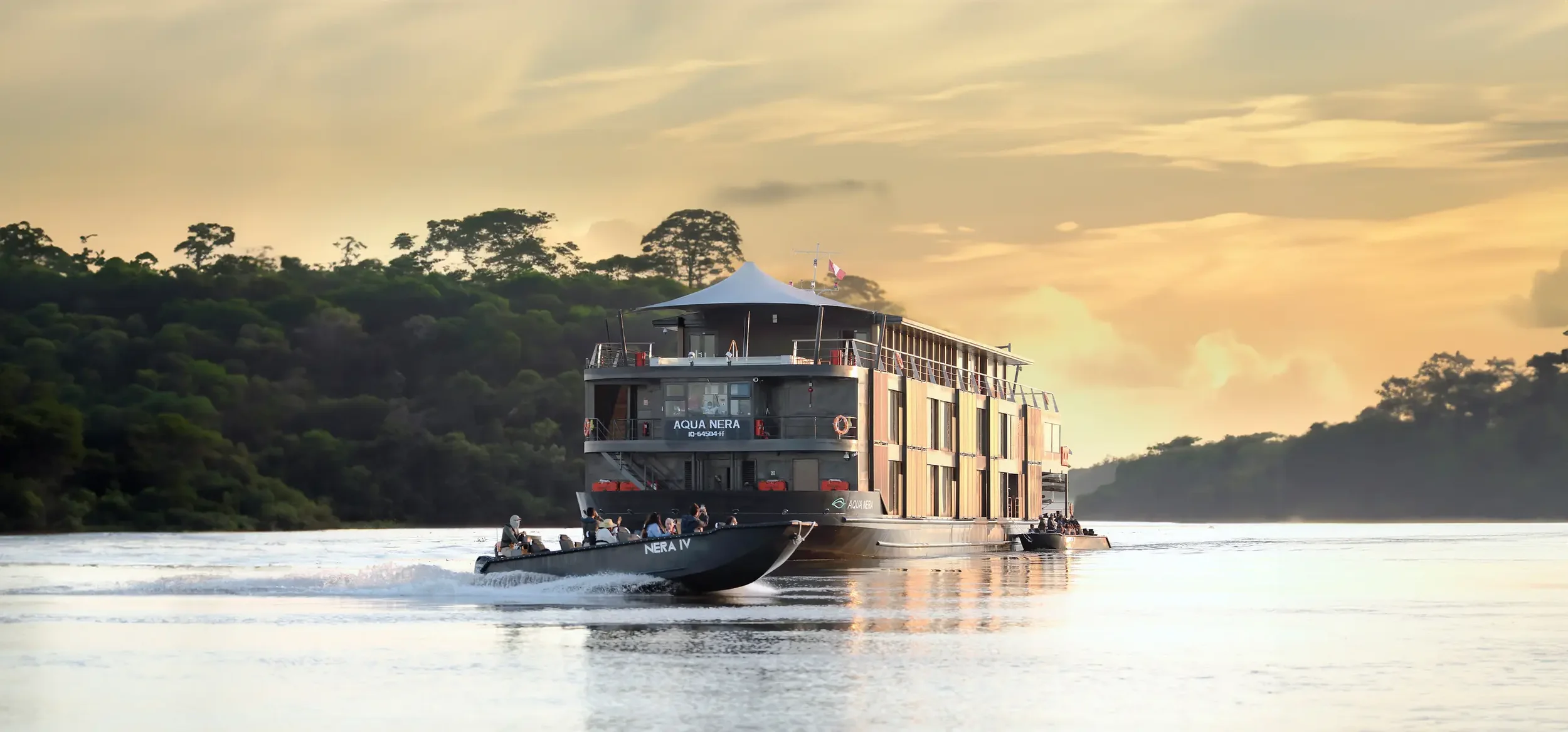 A large modern boat named Aqua Nera on a river at sunset, with a small motorboat passing by and lush green trees on the riverbank in the background.