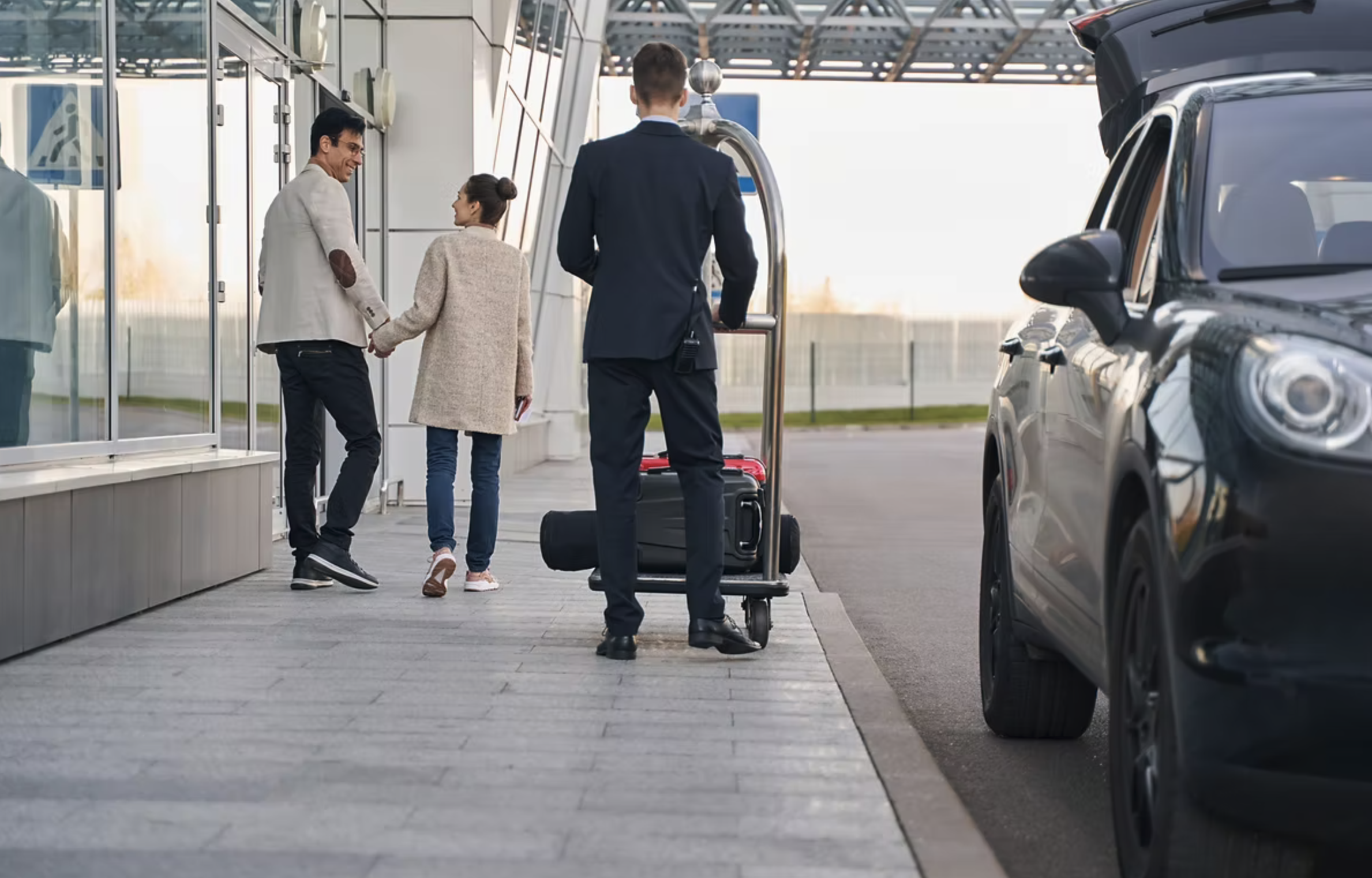 A man and a woman holding hands outside an airport terminal, with a luggage cart and a man in a suit nearby, and a black car parked to the side.