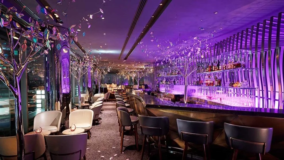 Elegant bar with purple lighting, modern decor, white and black chairs, decorative trees, and a glass railing