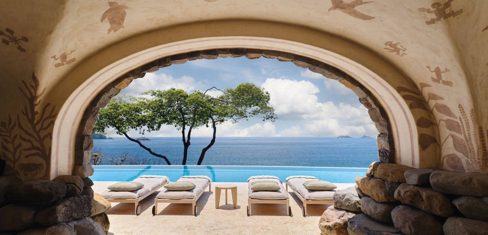 View of an ocean seen through an arched stone window with sun loungers and cushions on the terrace, and trees and islands in the distance.