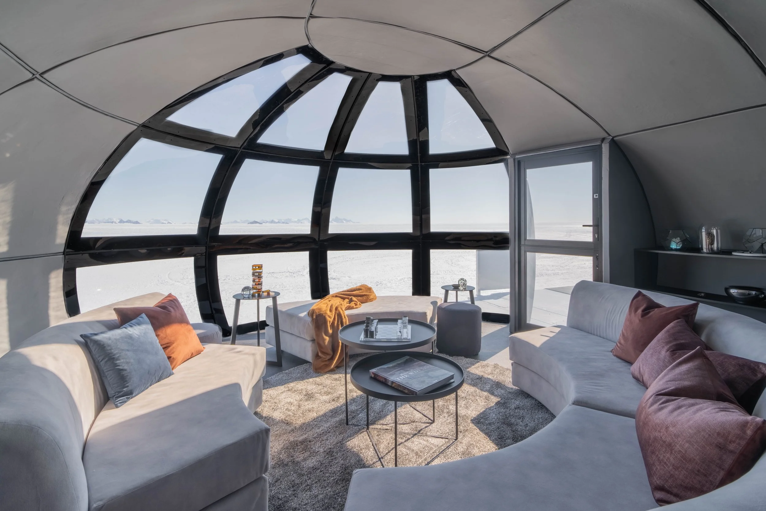 Interior of a modern, domed observation lounge with large arched windows offering a mountain and snow-covered landscape view, furnished with white curved sofas, colorful pillows, and round coffee tables.