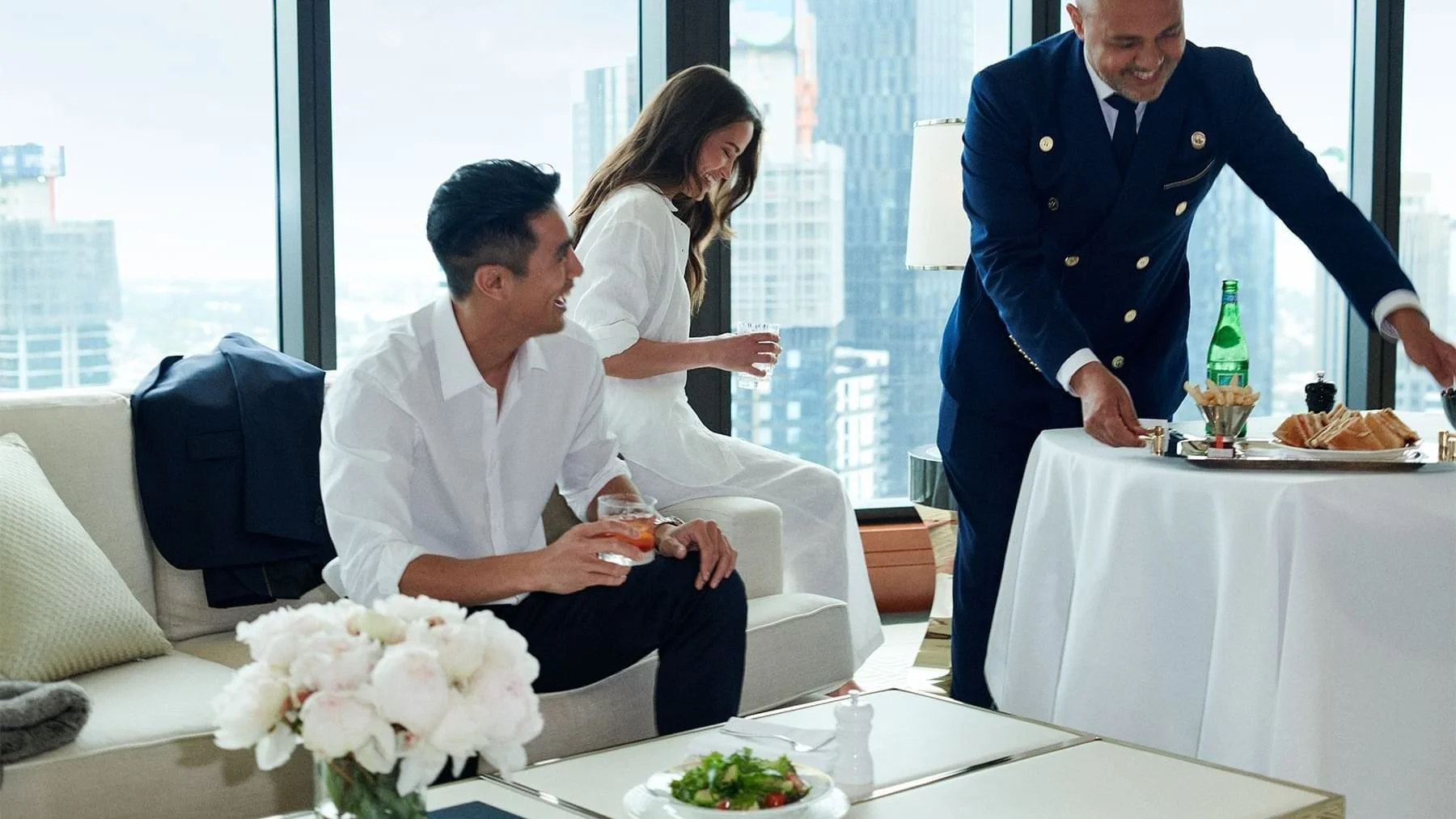 People enjoying a social event in a high-rise apartment with floor-to-ceiling windows, including a woman with brown hair in white dress, a man in white shirt holding a drink, and a man in a dark uniform serving hors d'oeuvres at a table.