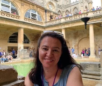 A woman smiling in front of a historic fountain at a tourist site with architecture and people in the background.