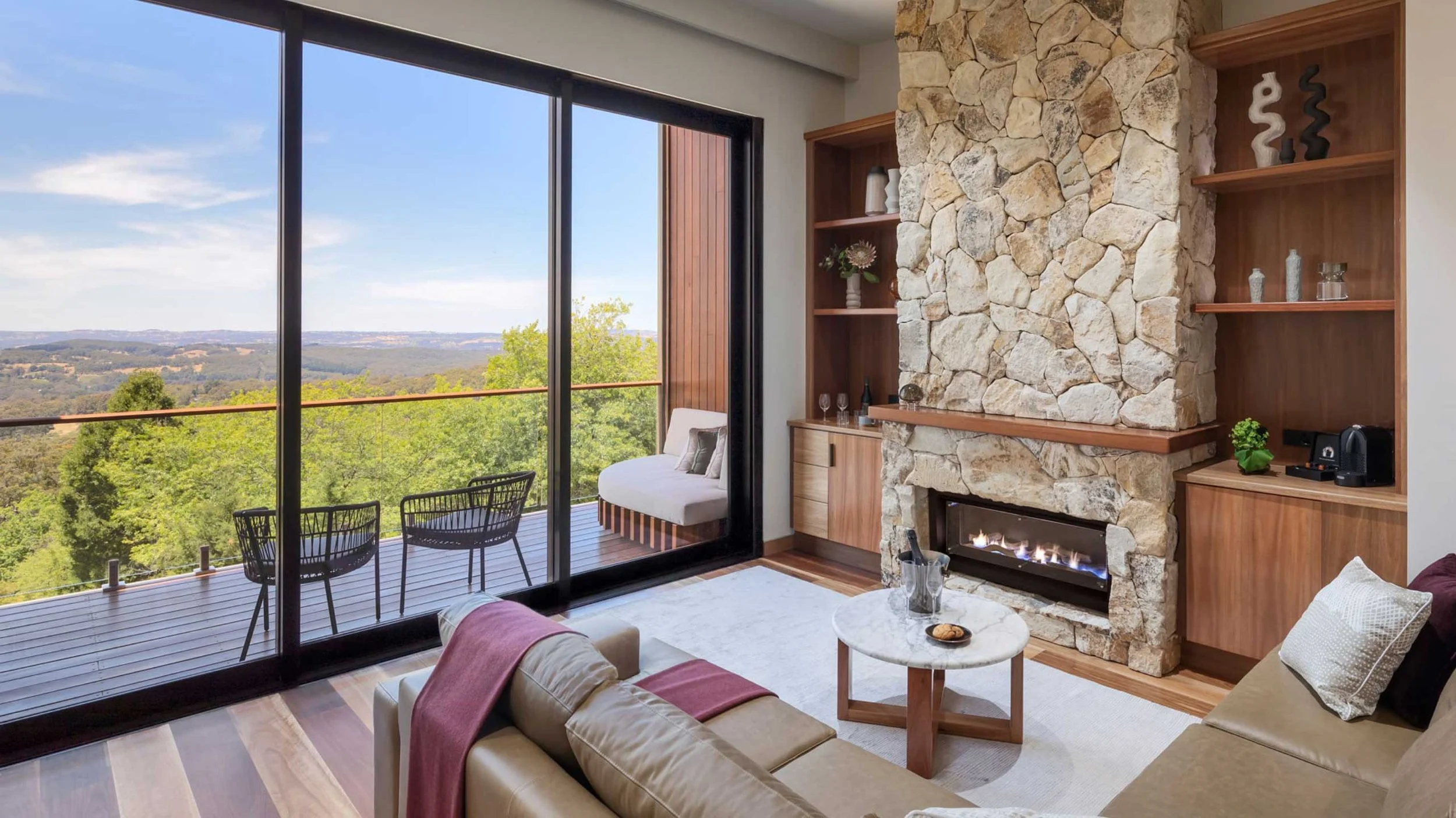 Living room with large sliding glass door leading to a balcony with outdoor chairs and a scenic view of green trees and distant hills. Inside, there is a beige sofa with a pink throw blanket, a round marble coffee table, and a stone fireplace with wooden shelves holding decorative items.