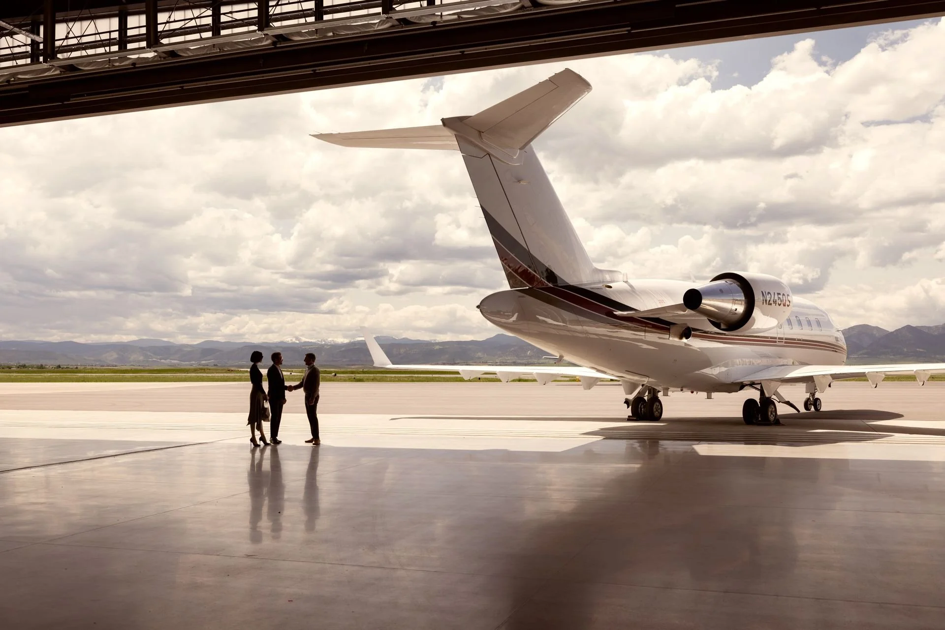 Three people in business attire shaking hands near a private jet inside an aircraft hangar, with the tarmac and mountains visible outside under partly cloudy skies.