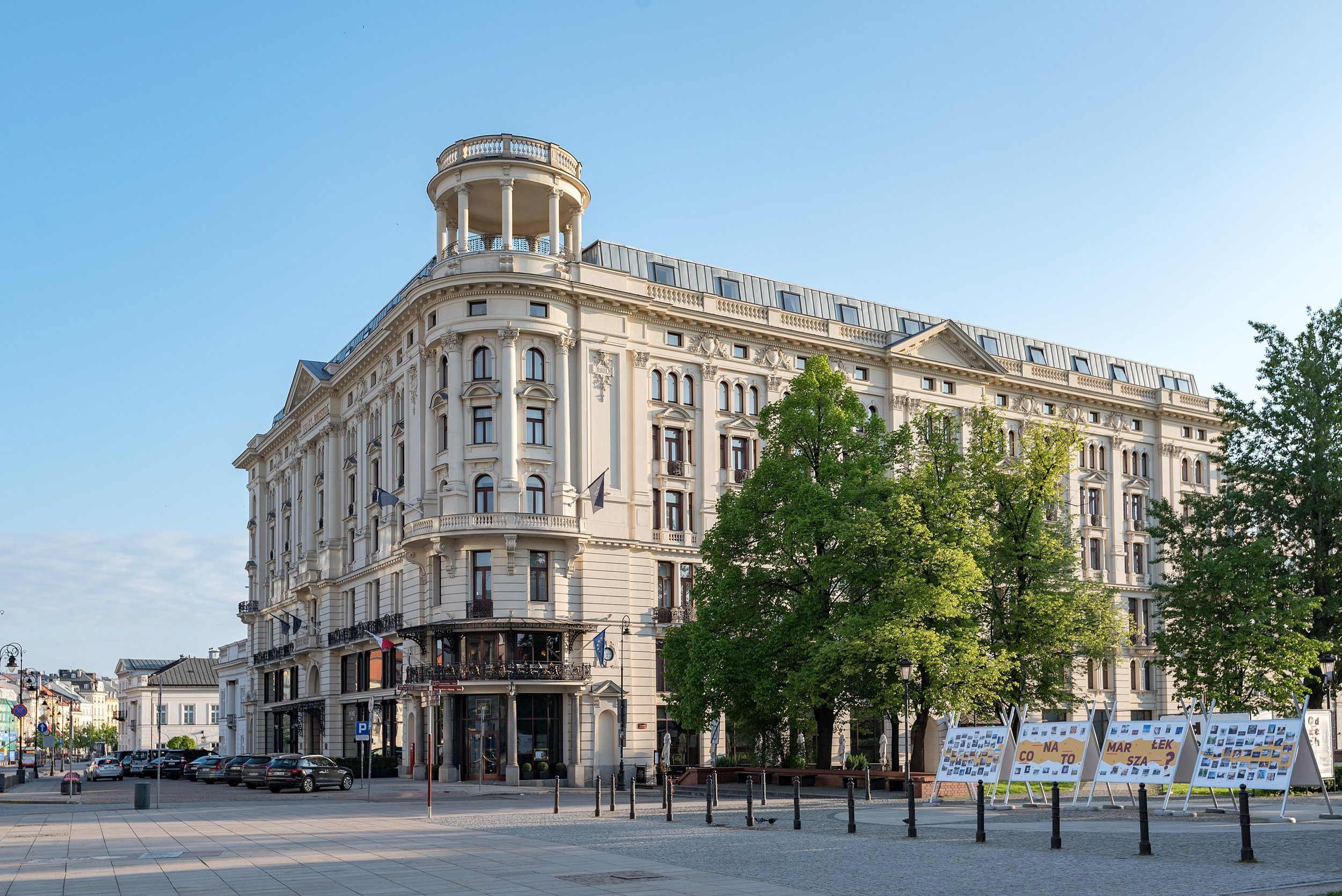 A large, ornate, white multi-story building with classical architectural details, surrounded by green trees and a city square with display boards and parked cars.