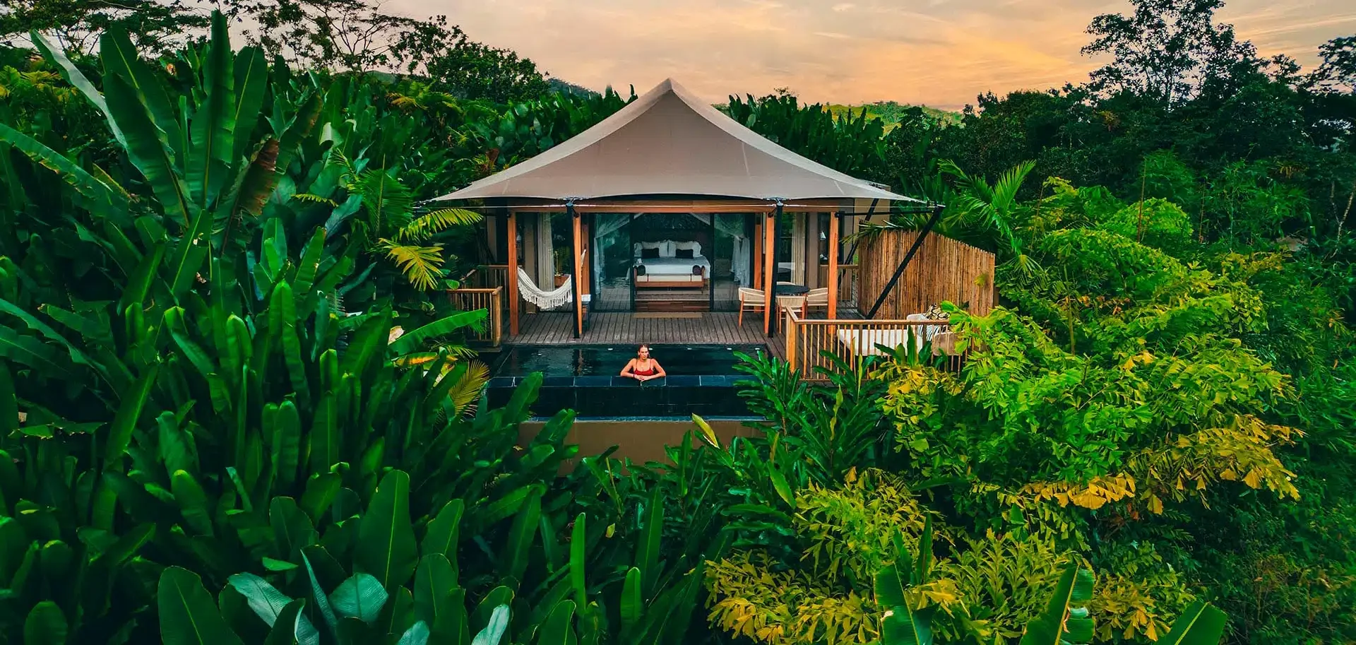 A person relaxing in a pool outside a wooden cabana surrounded by lush green tropical foliage at sunset.