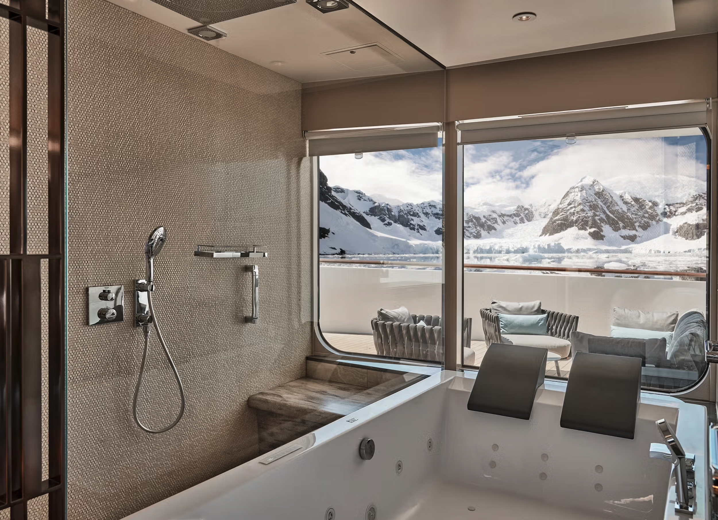 A luxurious bathroom with a large bathtub and a picturesque view of snow-covered mountains through the glass window, with outdoor seating on a balcony.