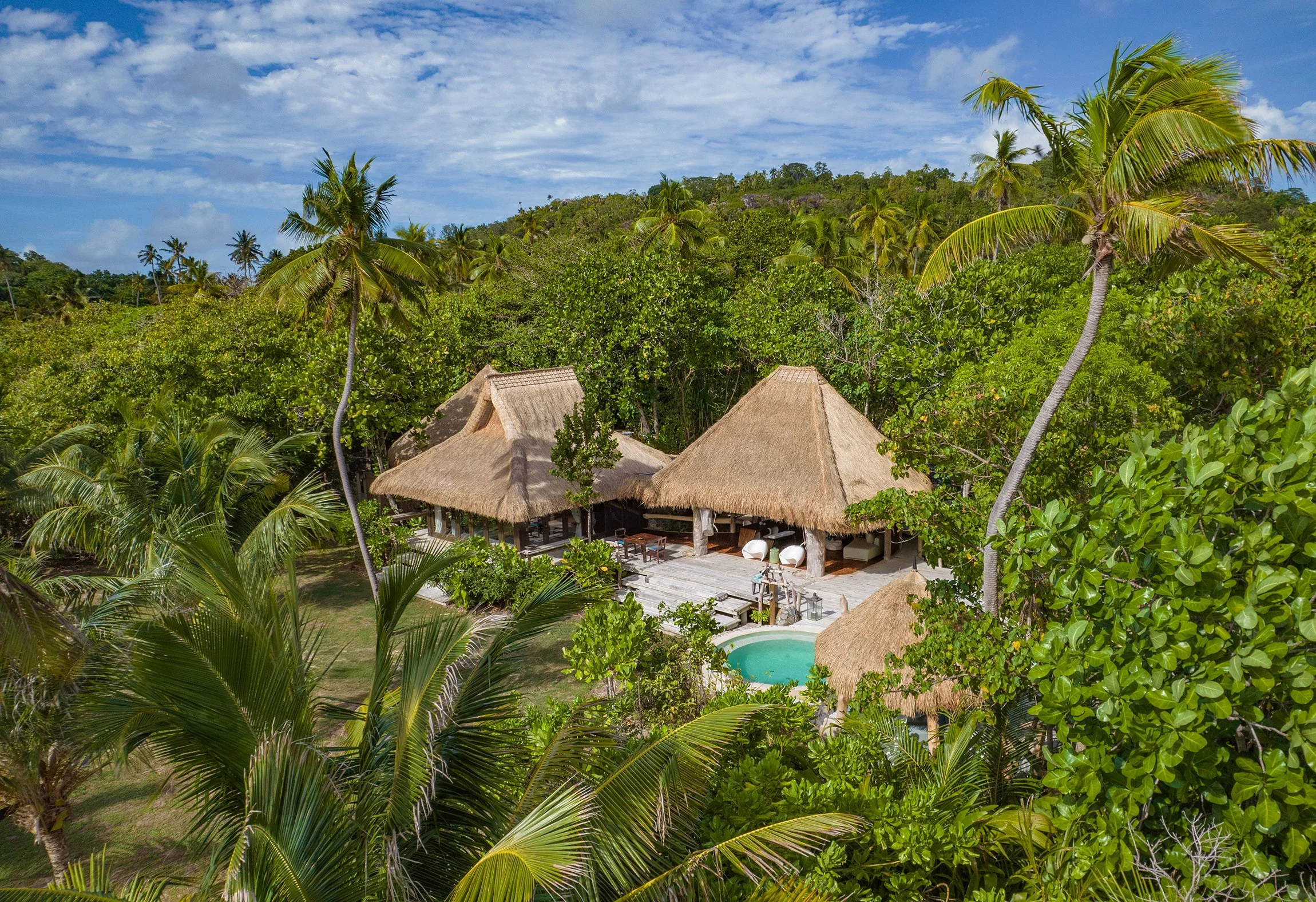 A tropical resort with thatched roof buildings, surrounded by lush green palm trees and foliage, and a small swimming pool, under a blue sky with clouds.