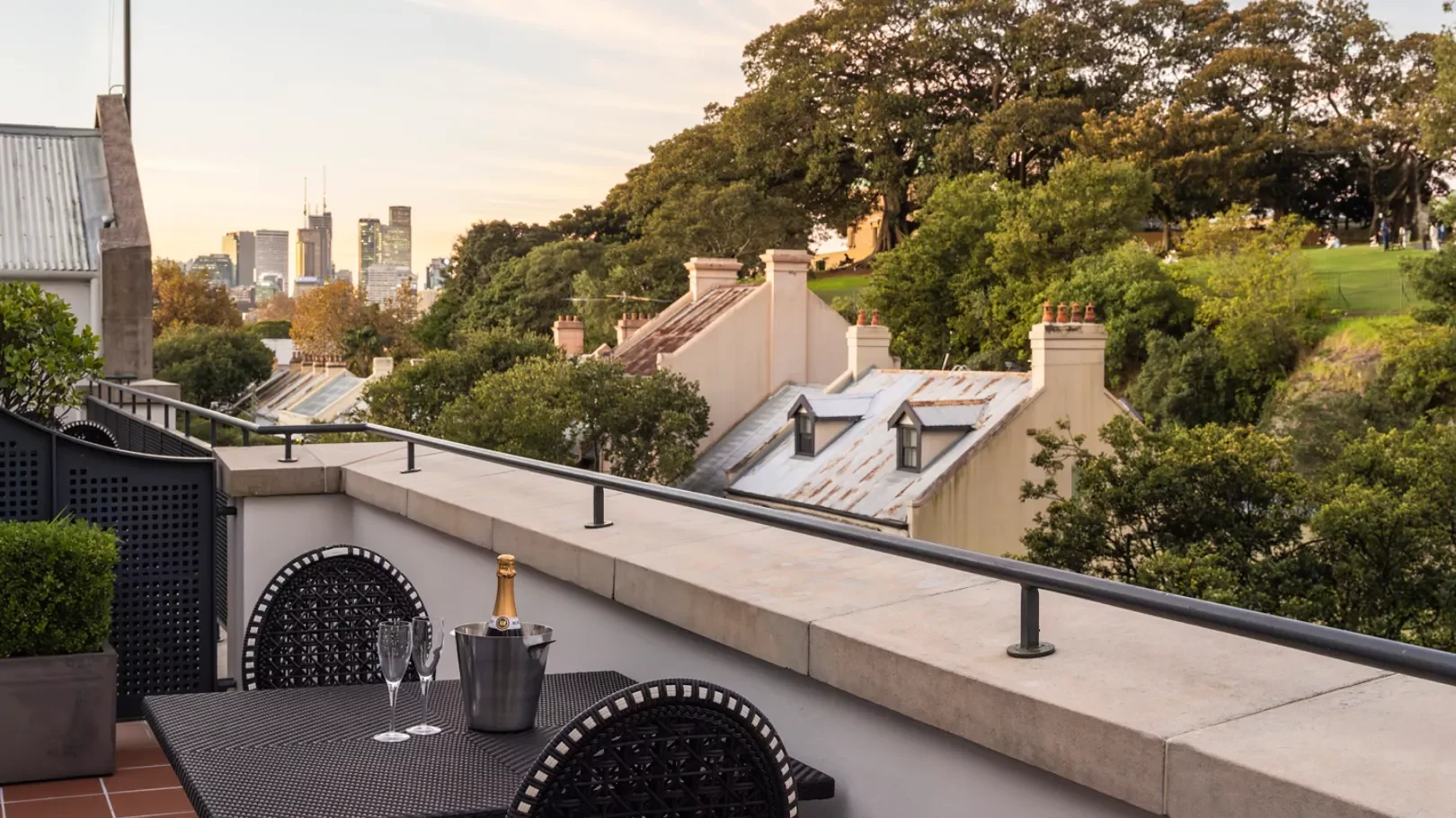 Urban rooftop terrace with table, chairs, and a bottle of champagne in an ice bucket, overlooking residential rooftops, trees, and a city skyline at sunset.