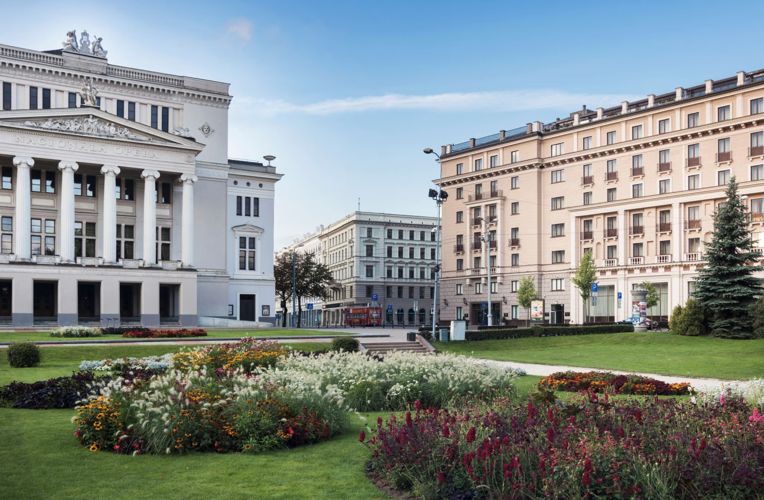 City park with flowering garden, historic buildings, and a red double-decker bus in the background.