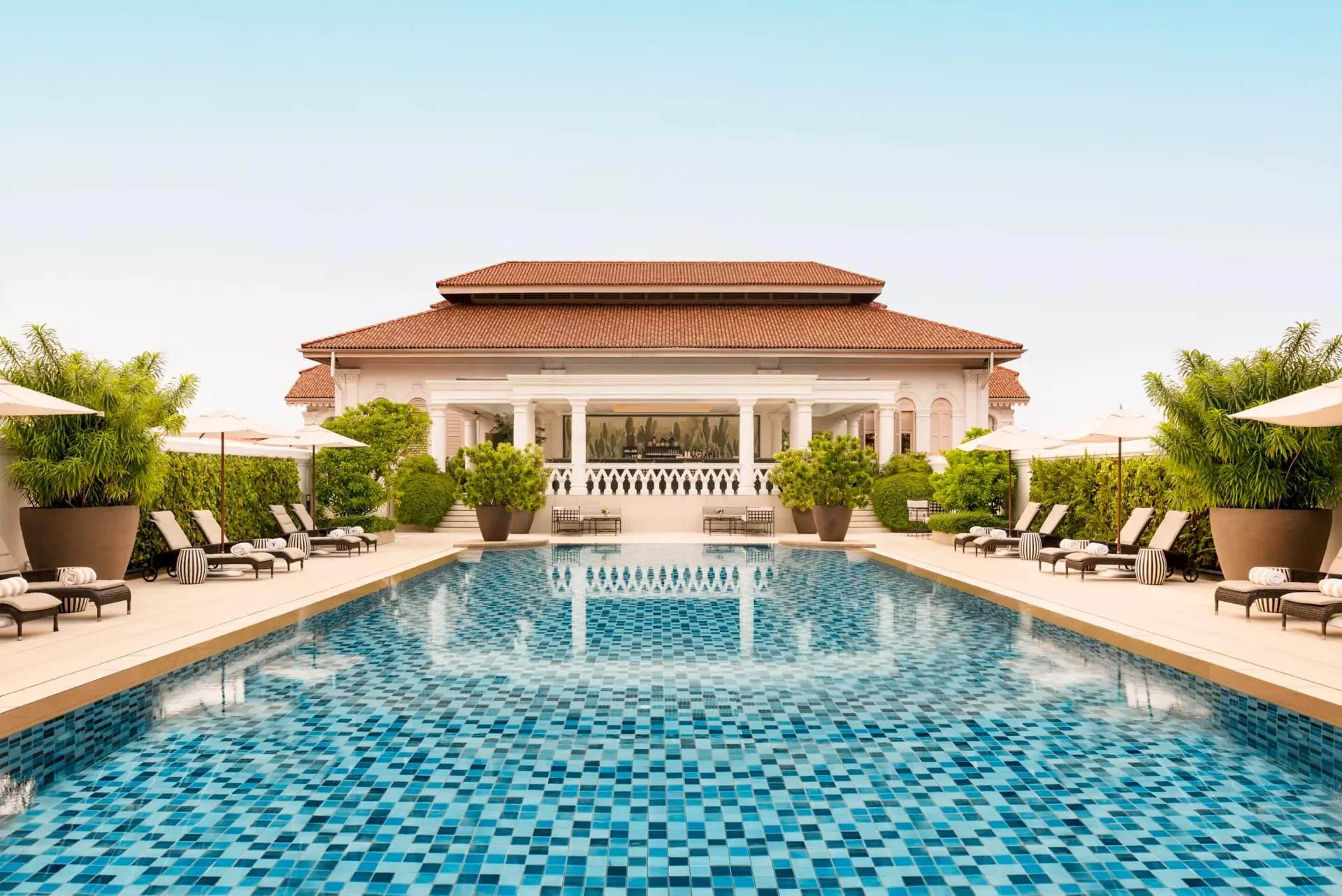 Luxury hotel pool area with lounge chairs, umbrellas, and lush green plants, overlooking a grand building with columns and a tiled roof.