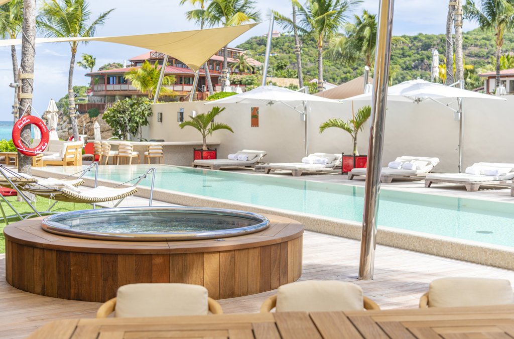 Poolside area at a vacation resort with lounge chairs, umbrellas, palm trees, and a hot tub.