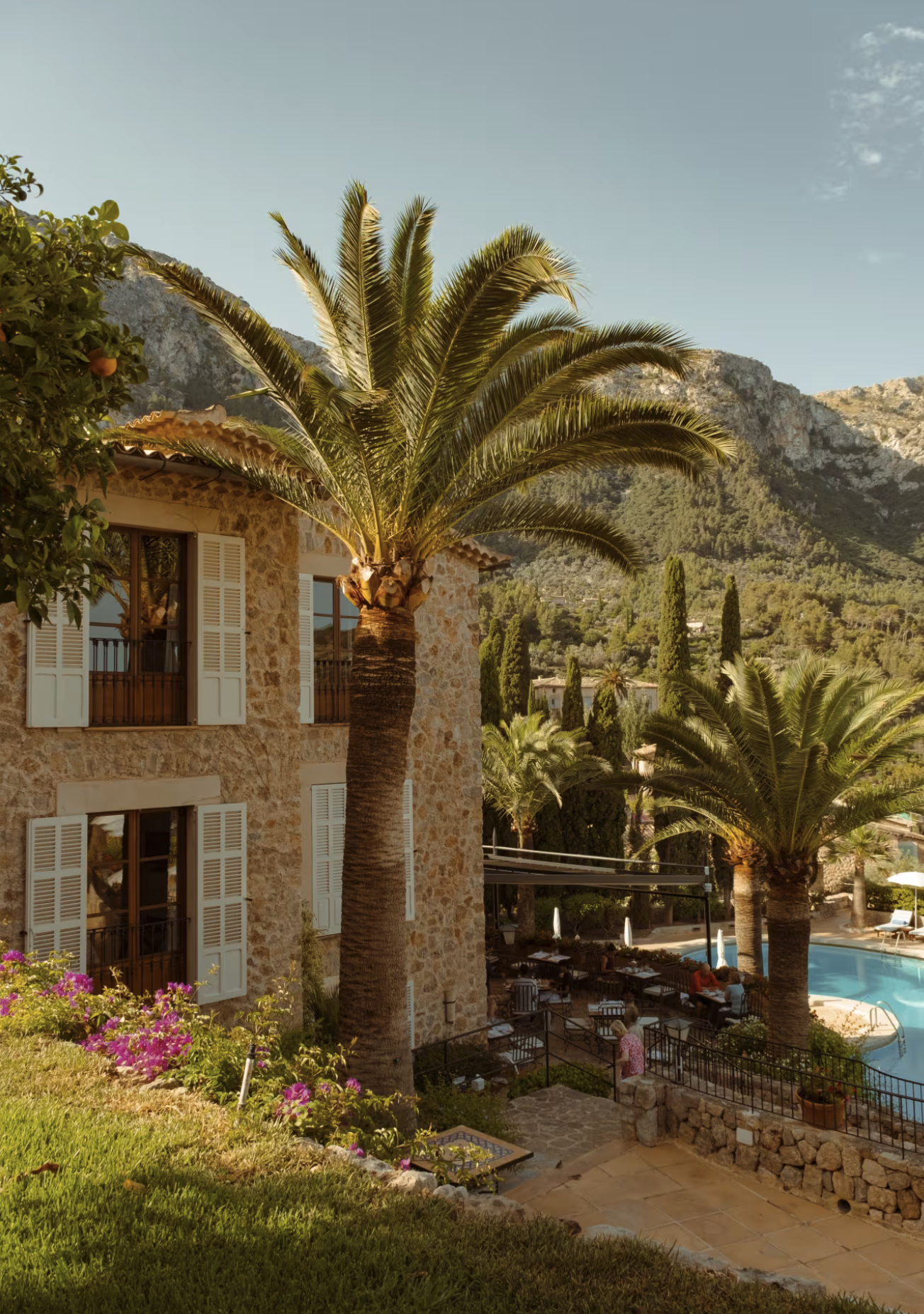 A stone building with white shutters, tall palm trees, a swimming pool, and outdoor seating area surrounded by lush greenery and mountains in the background.