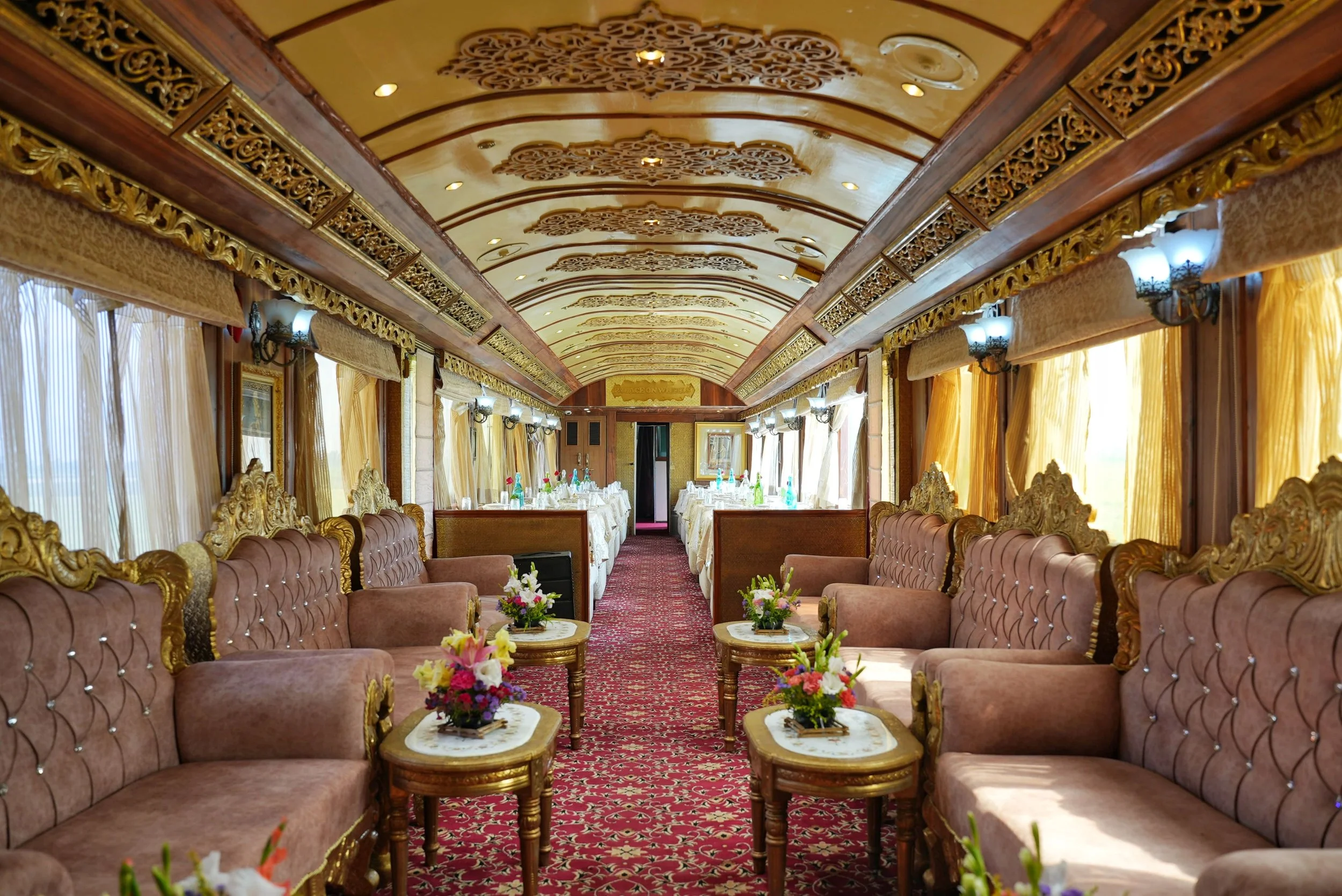 Elegant dining area inside a luxurious vintage train carriage with plush pink sofas, ornate gold detailing, small floral arrangements on tables, and long banquet tables set with white linens and glassware at the far end