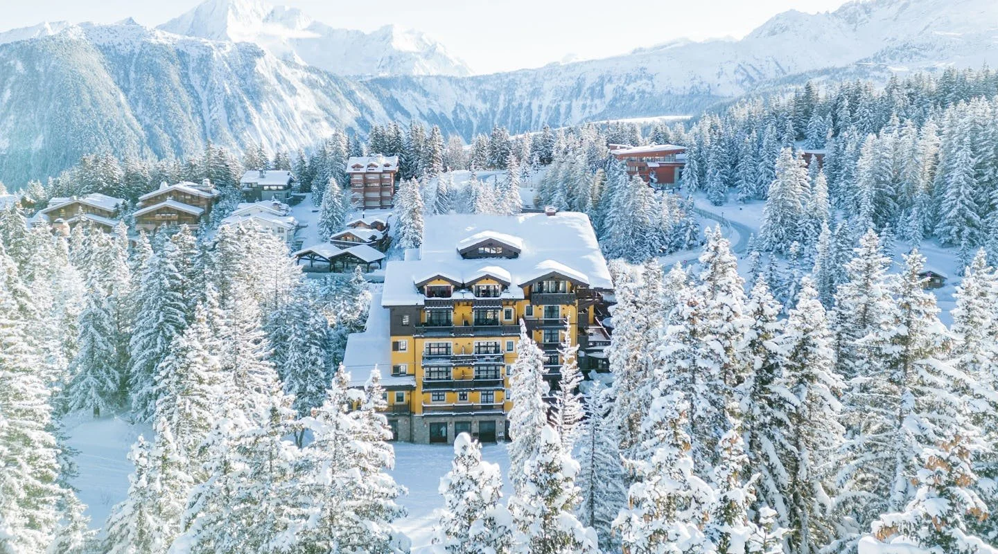 Snow-covered mountain landscape with numerous chalet-style buildings and a large yellow apartment complex surrounded by snow-laden pine trees.