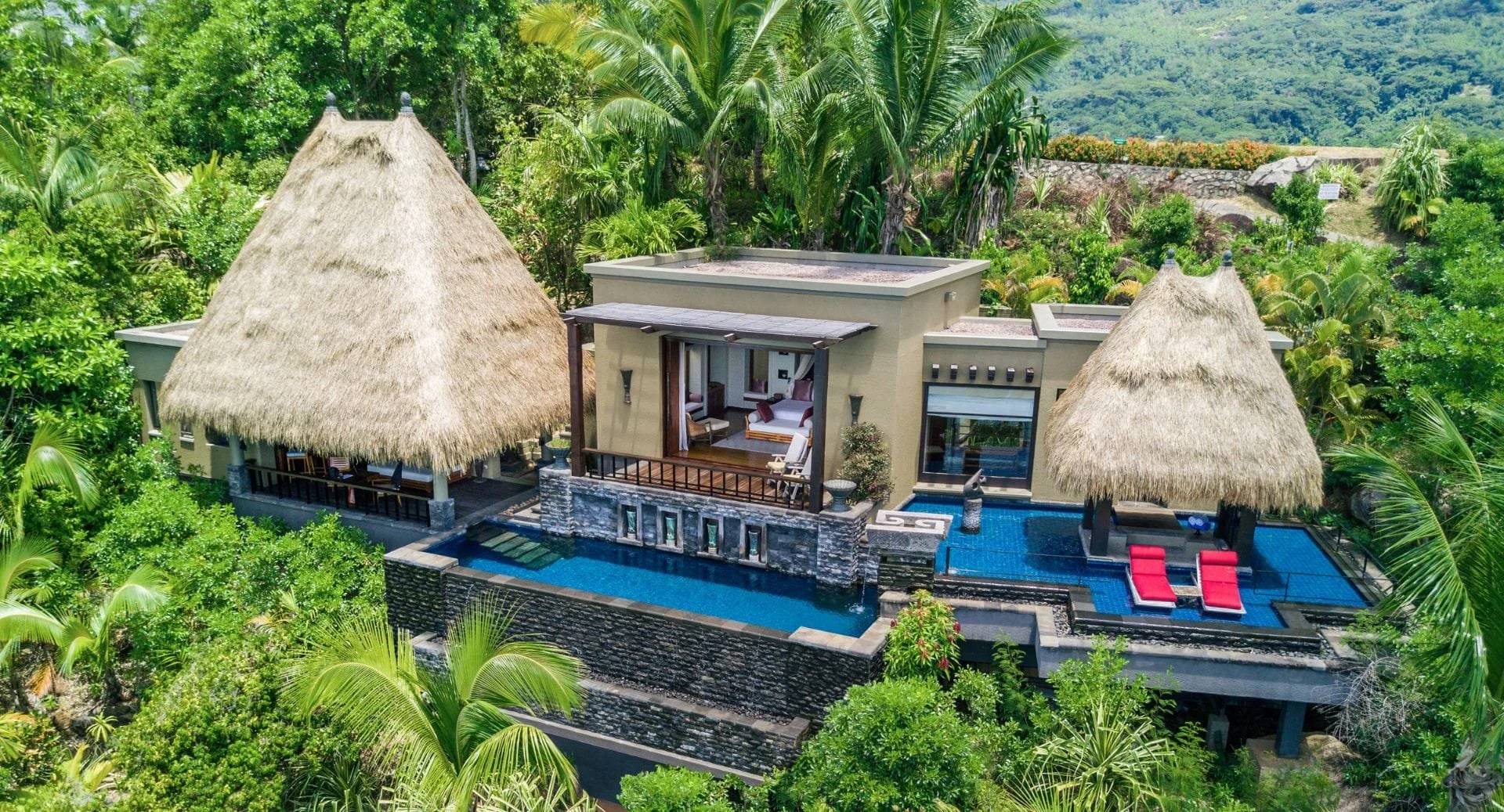 Luxury tropical villa surrounded by lush green foliage, featuring a thatched roof hut, modern building with large windows, and a swimming pool with red lounge chairs.
