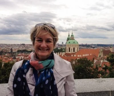 Smiling woman with short blonde hair wearing sunglasses, a light jacket, and a colorful scarf, standing in front of a cityscape with a historic church and buildings under cloudy sky.