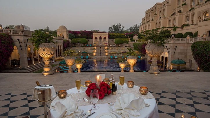 A luxurious outdoor dining setup overlooking a lavish palace-style building and water feature with decorative urns and greenery at dusk.