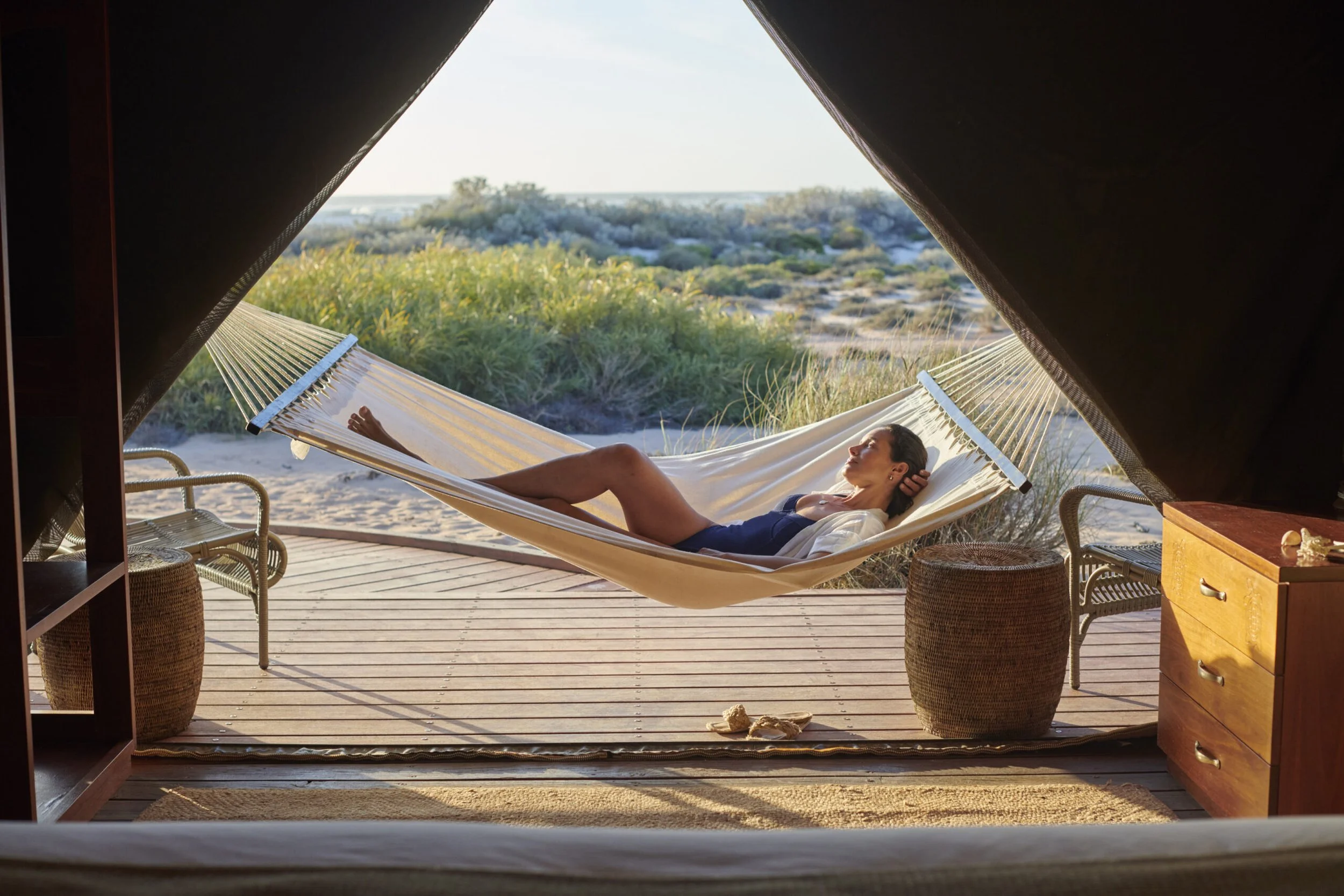 Woman relaxing on a hammock outside a tent overlooking a sandy beach and green dunes during sunset.