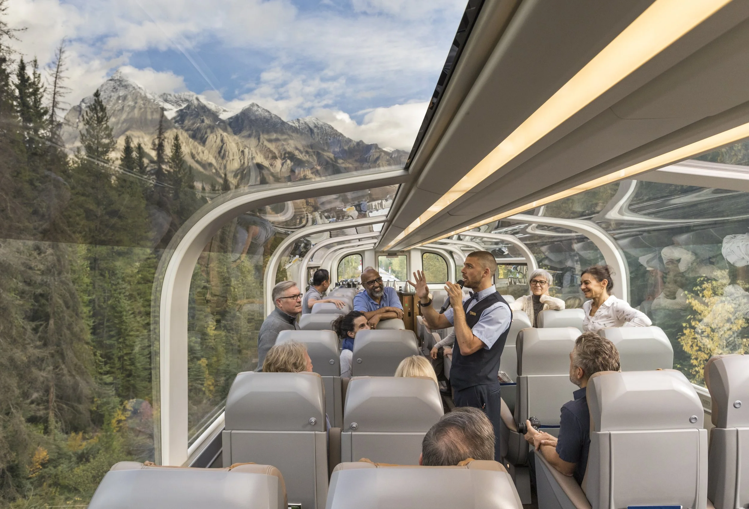 Inside a glass tunnel on a train, with scenic mountain and forest views outside. A man stands and gestures as he talks to a group of diverse passengers seated around him.