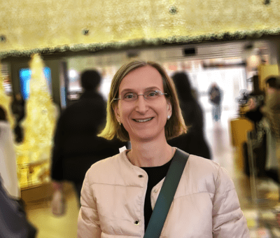 Woman with glasses smiling in an indoor shopping or airport terminal with people in the background.