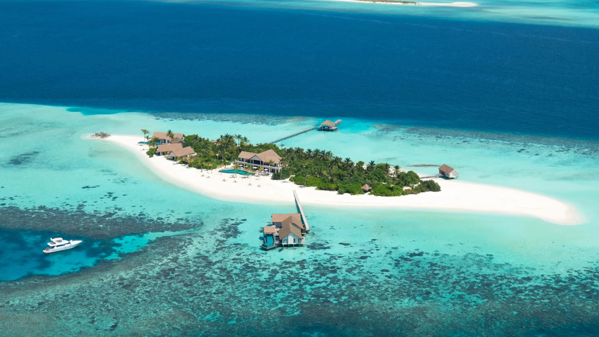 Aerial view of a small island with white sandy beaches, surrounded by turquoise water and coral reefs, with several overwater bavilions connected by boardwalks, and a boat anchored nearby.