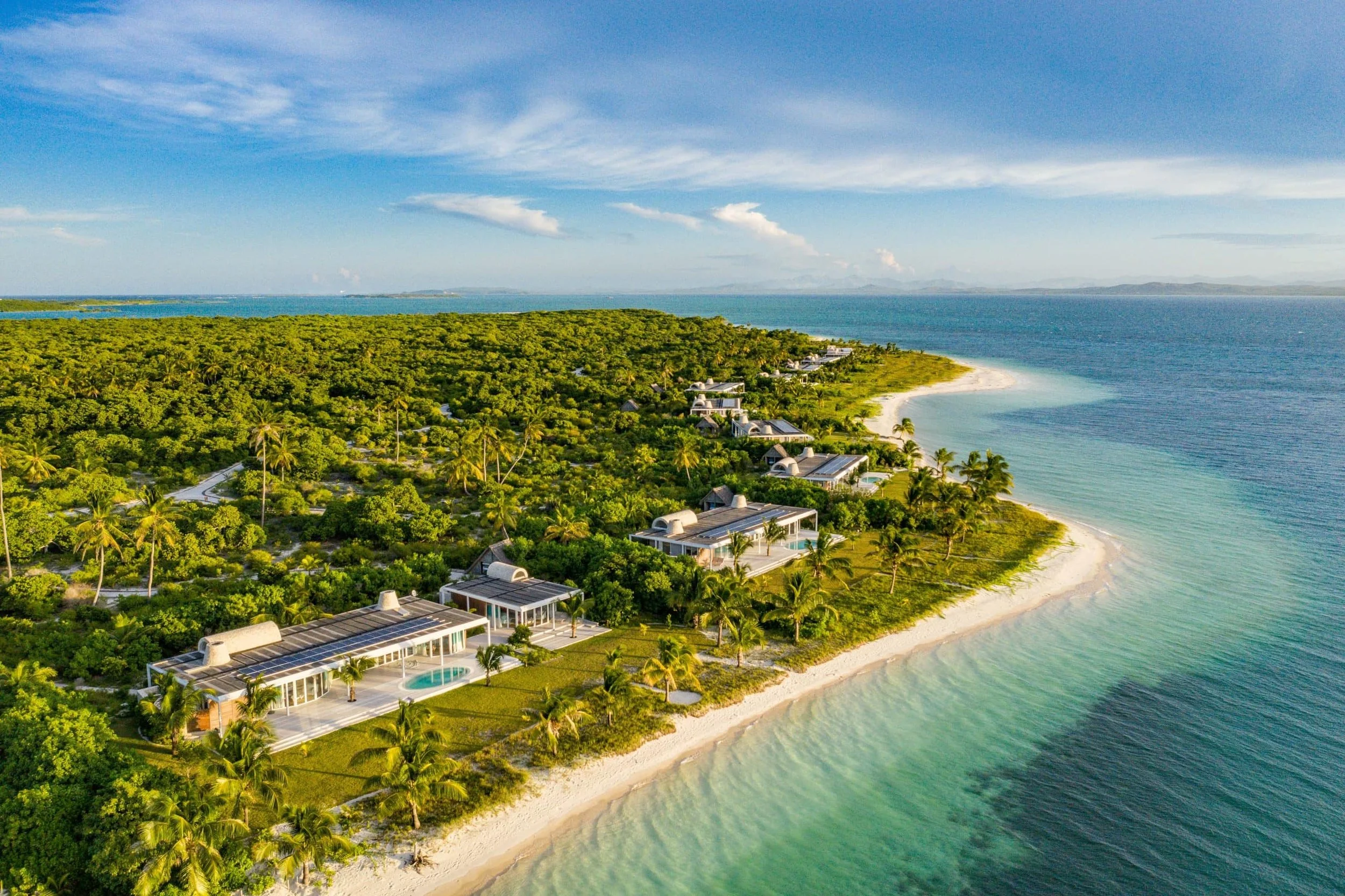 Aerial view of a beachfront resort with modern villas surrounded by lush greenery and palm trees, overlooking clear blue ocean waters with sandy shores.