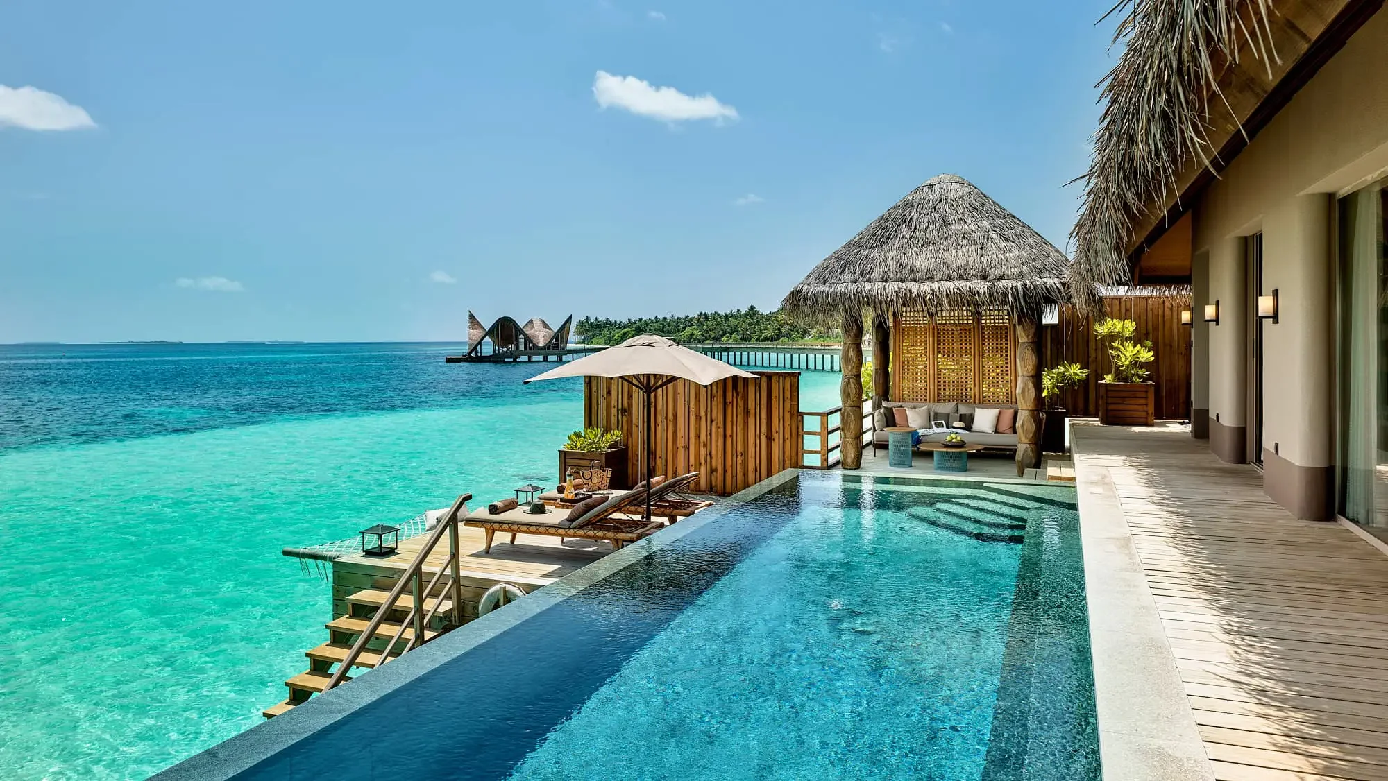 Luxury overwater bungalow with private infinity pool, deck chairs, and shaded seating area overlooking turquoise ocean with a distant island and modern building in the background.
