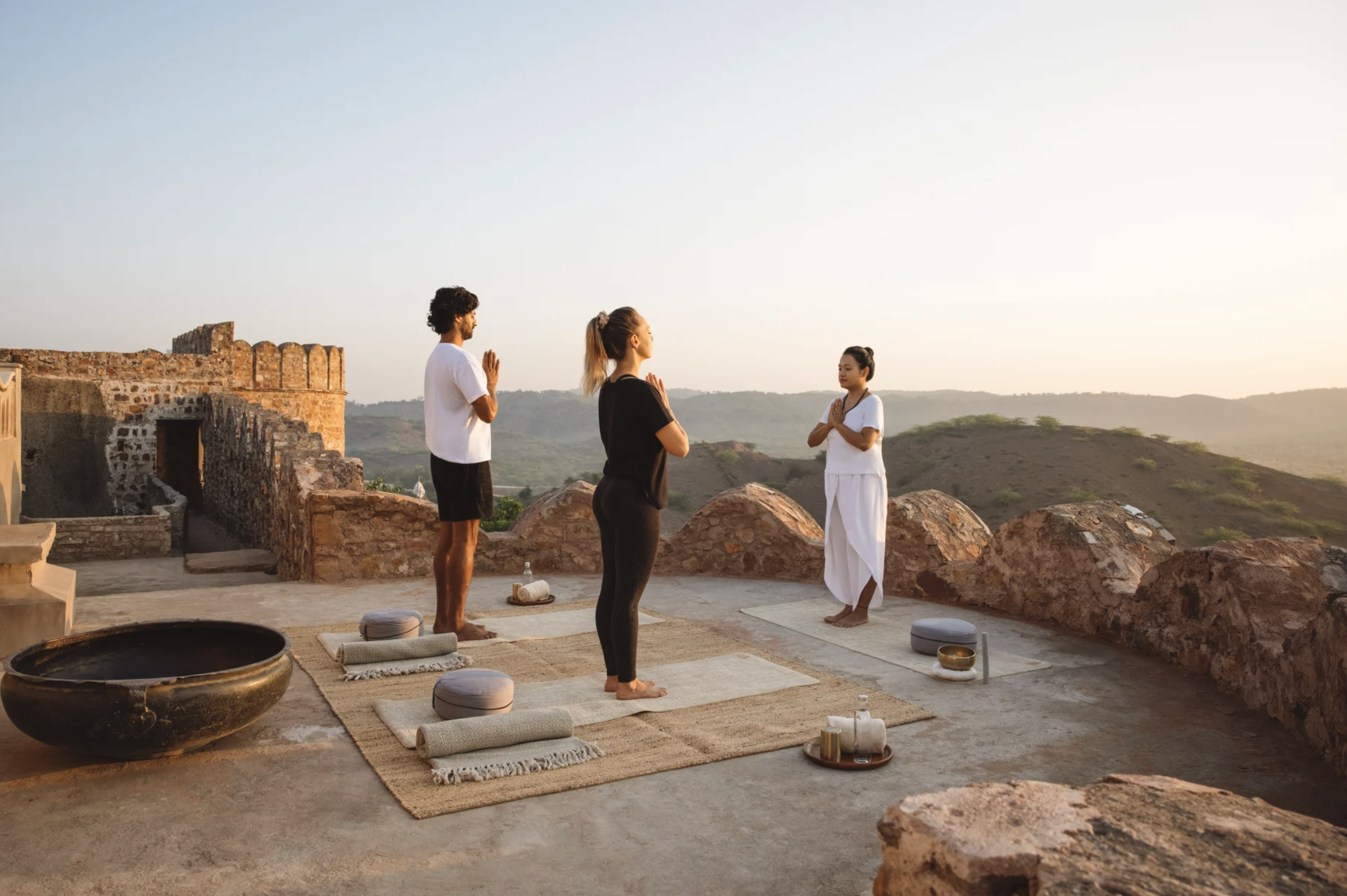 Three people practicing yoga on a rooftop terrace overlooking hills at sunrise or sunset, with stone walls and yoga mats.