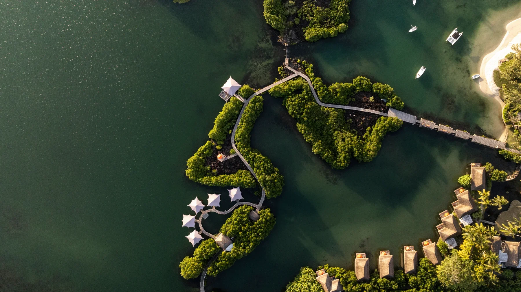Aerial view of a park with walkways on small islands surrounded by water, with boats anchored nearby and trees along the shoreline and in the islands, some structures and umbrellas visible.