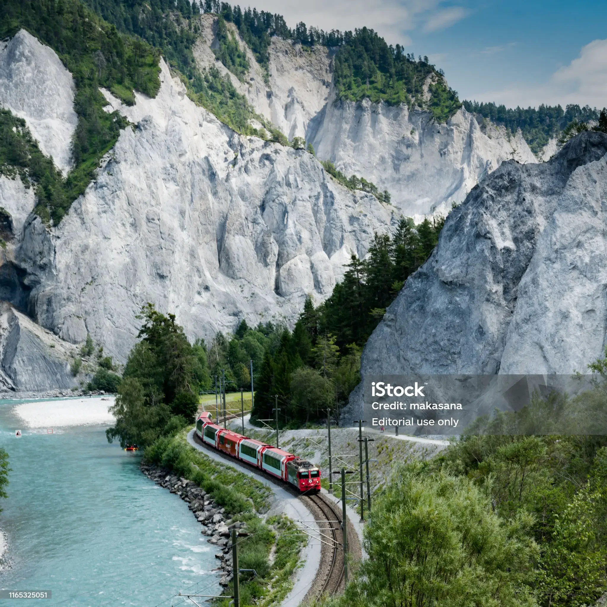 A train traveling along a railway track beside a turquoise river, with tall white and gray mountains and green trees in the background.