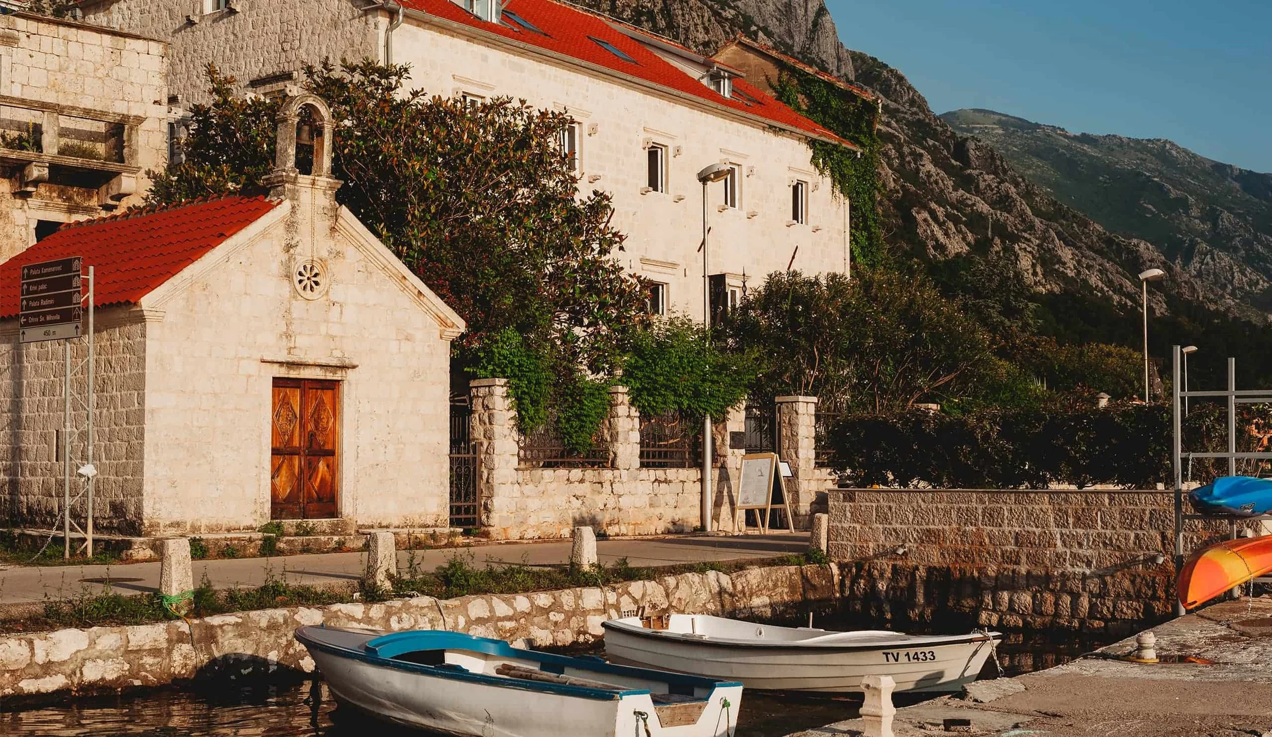 A small harbor with two boats docked, a stone building with a red tile roof, trees, a stone wall, and mountains in the background.