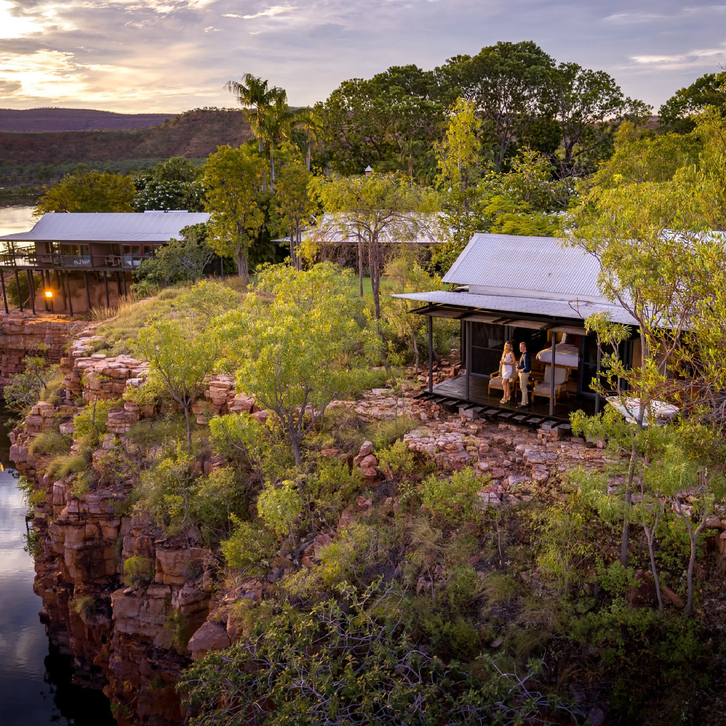 View of two modern houses on a rocky cliffside surrounded by trees, with a river and hills in the background during sunset.