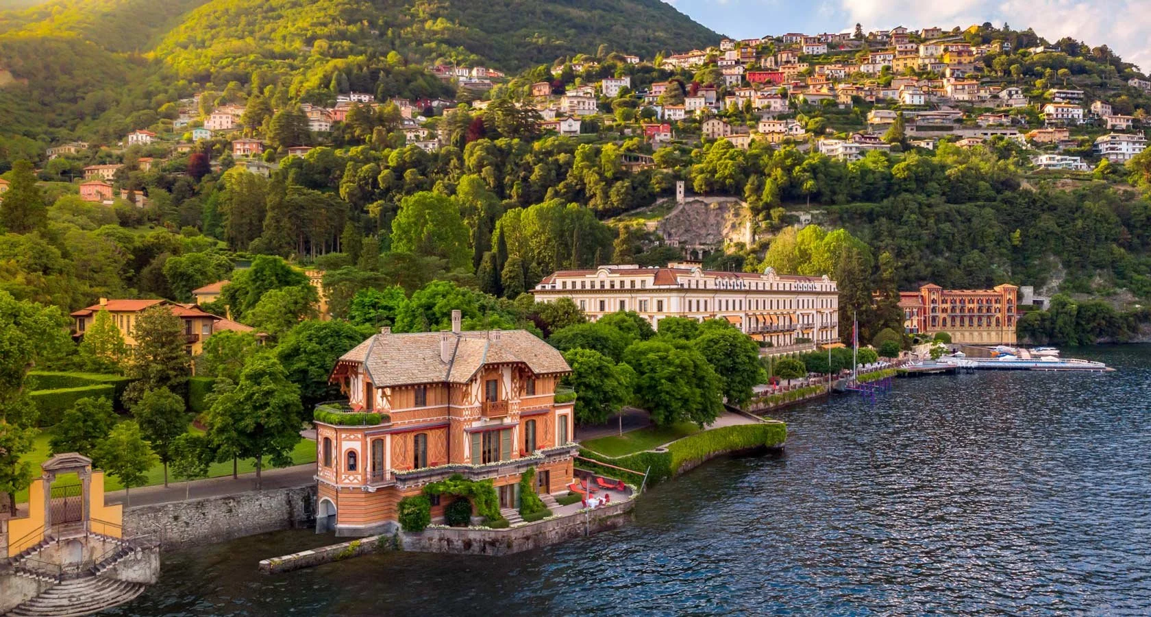 Lakeside view of a hillside town with colorful houses, lush green trees, and a large historic building near the water, with boats docked at a pier.