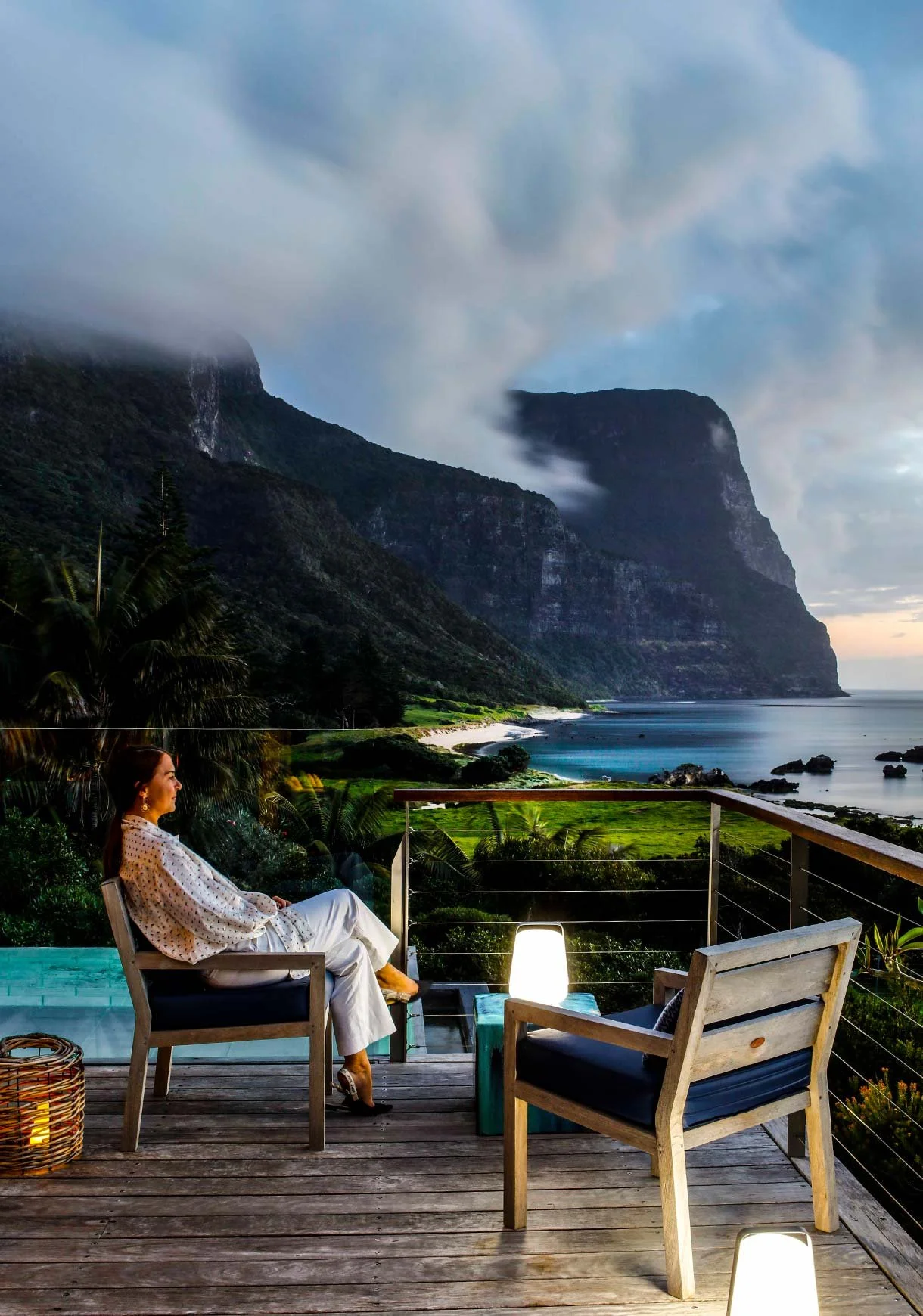 A woman sitting on a deck chair on a balcony overlooking a scenic coastal landscape with mountains, ocean, and lush greenery during dusk or early evening.