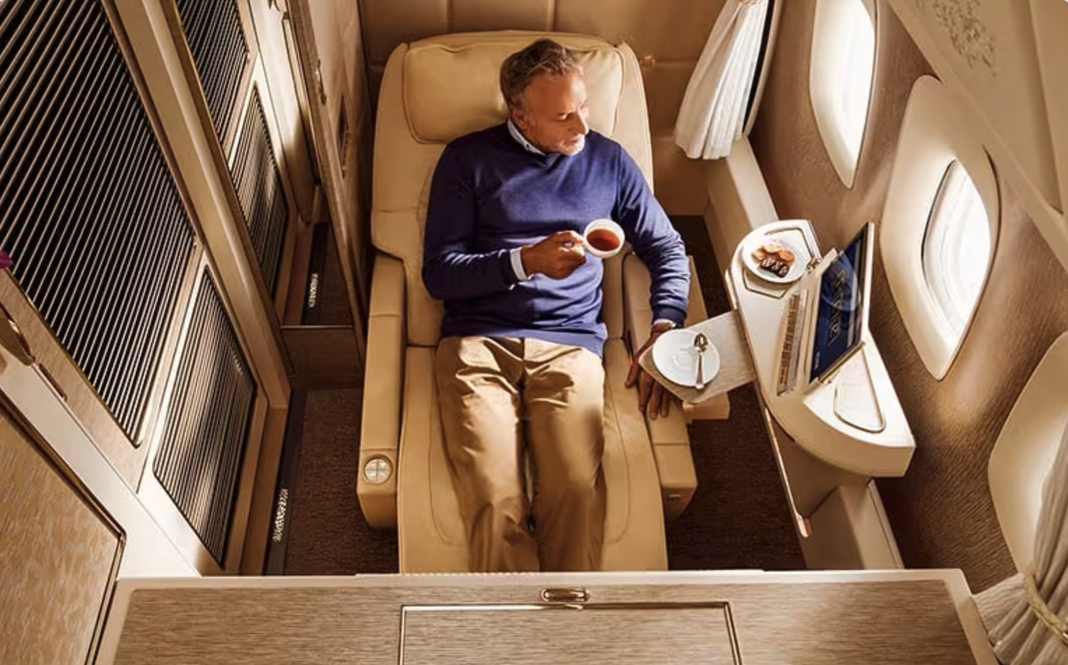 A man relaxing on a luxurious airplane seat, holding a cup of tea or coffee, with a laptop, a plate of cookies, and a spoon on a tray table beside him, surrounded by windows and curtains.