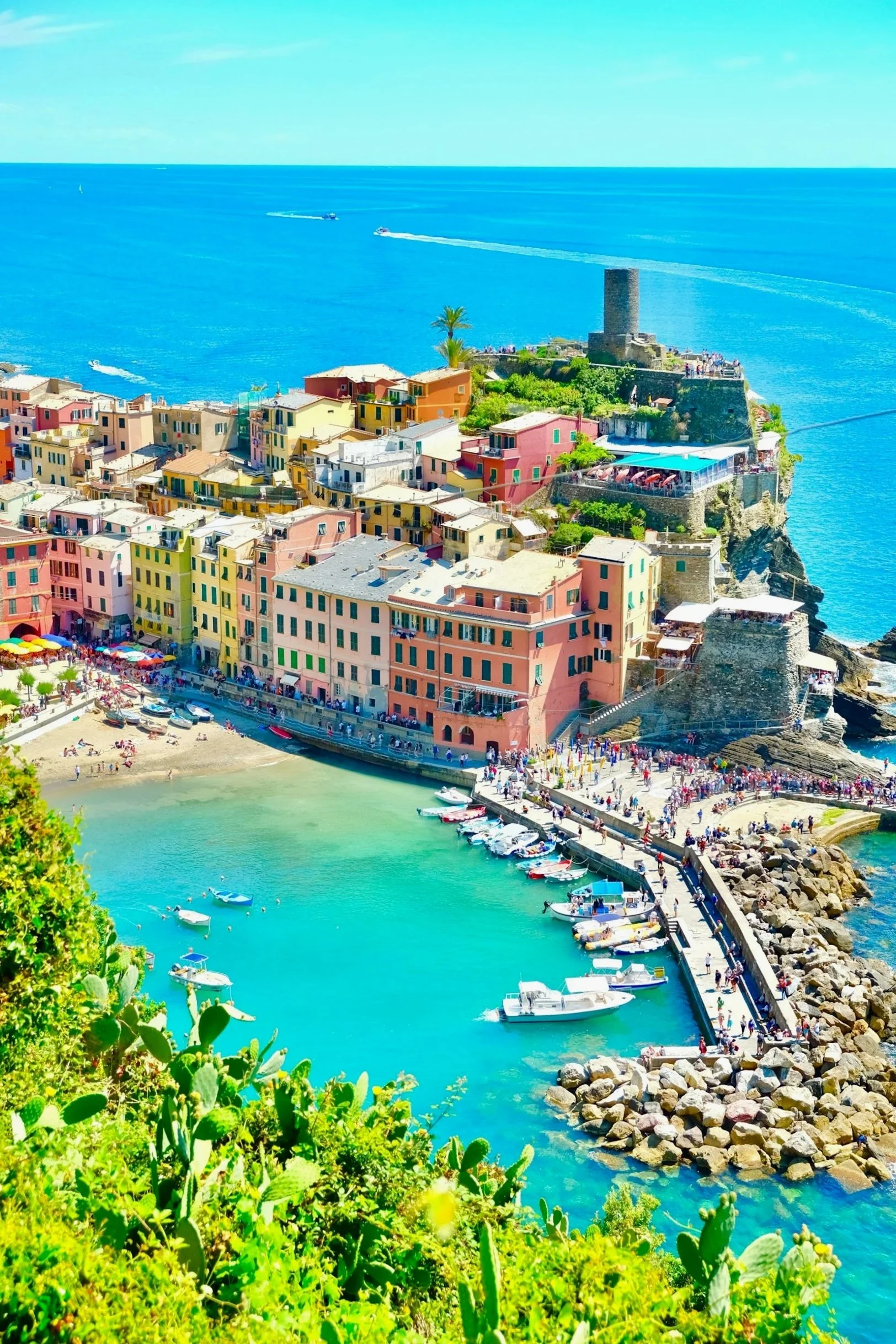 Colorful seaside village on a rugged coast with boats docked in turquoise water and a tower on a cliff overlooking the ocean.