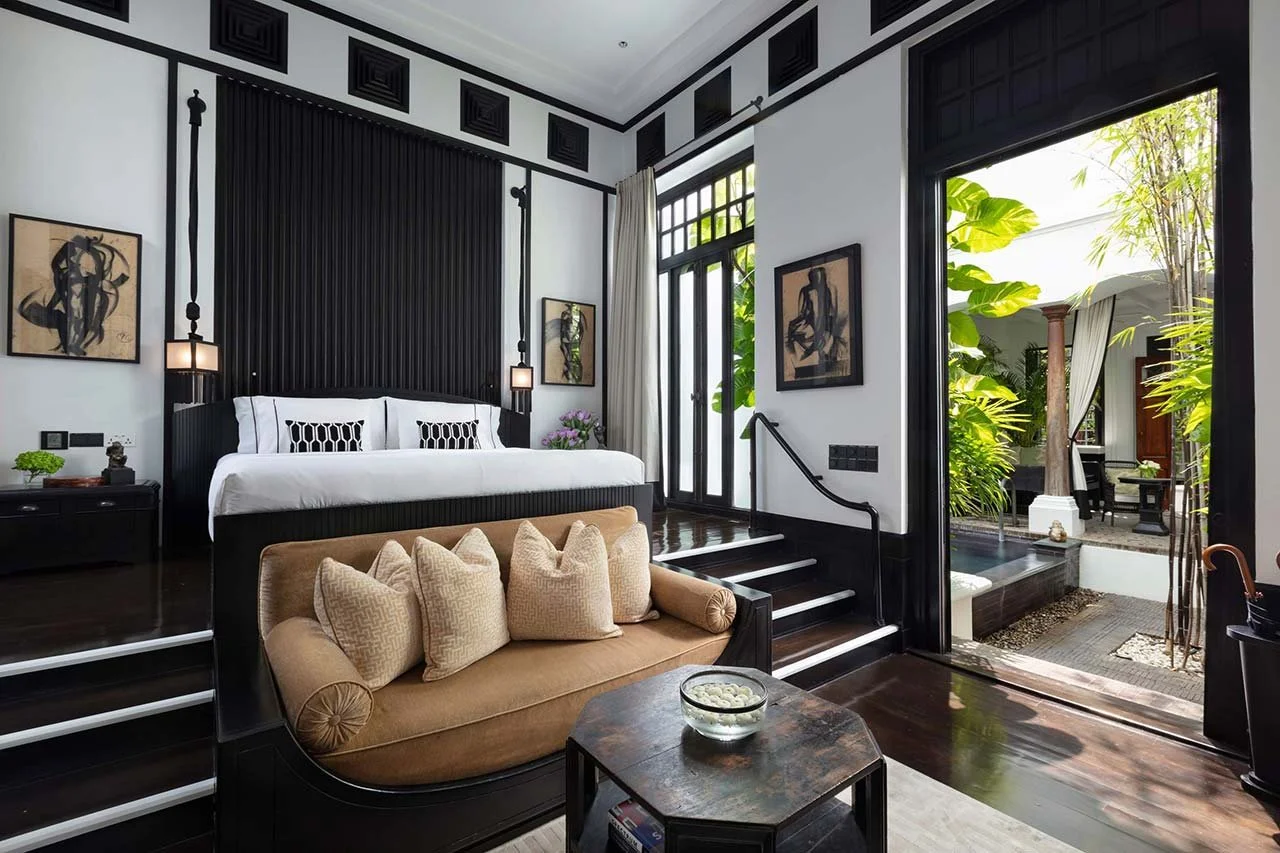 Luxurious bedroom with black and white decor, featuring a bed with white bedding, a beige sofa with cushions, and large windows with lush greenery outside.