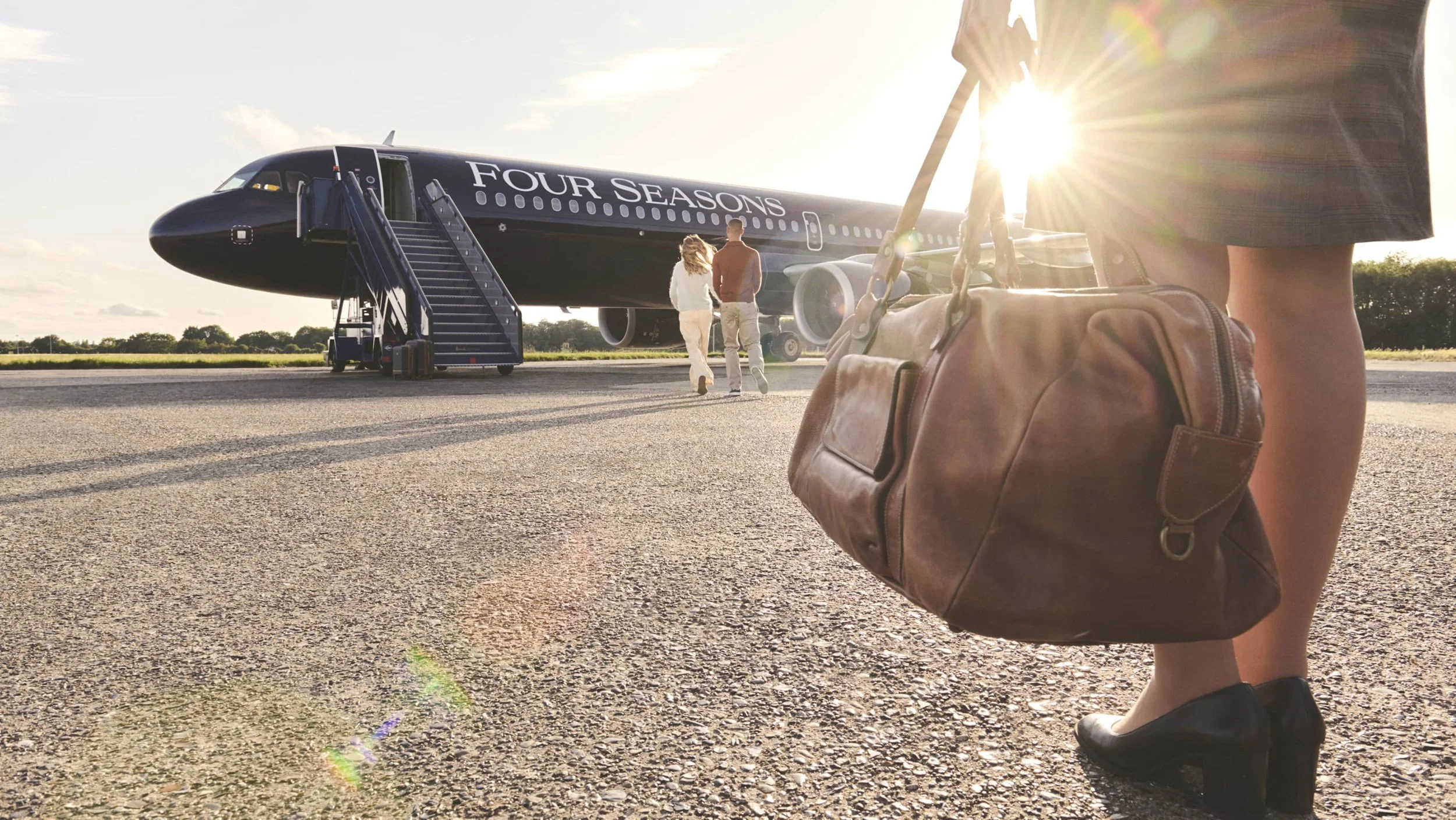 A woman holding a brown handbag in front of an airplane with the logo "Four Seasons" on the side, with two people walking towards the plane, and the sun shining brightly behind them.