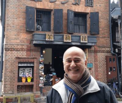 Smiling man standing outside a brick building with black shutters and a sign above the entrance that reads 'Bar & Grill' in Chinese characters.