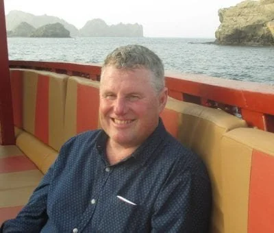 A smiling man on a boat with a scenic water and rocky island background.