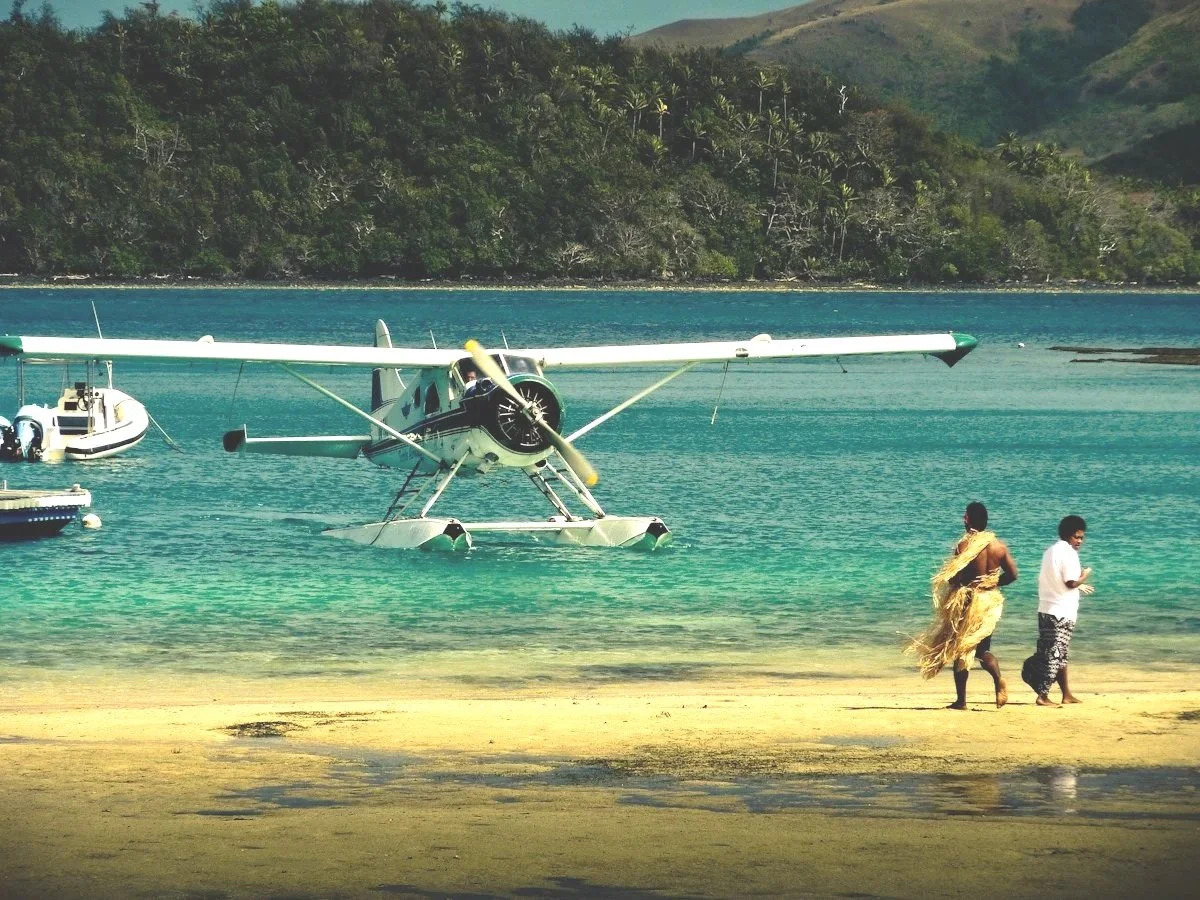 Seaplane on turquoise water near a sandy beach with two men walking, one in traditional attire, with lush green hills in the background.
