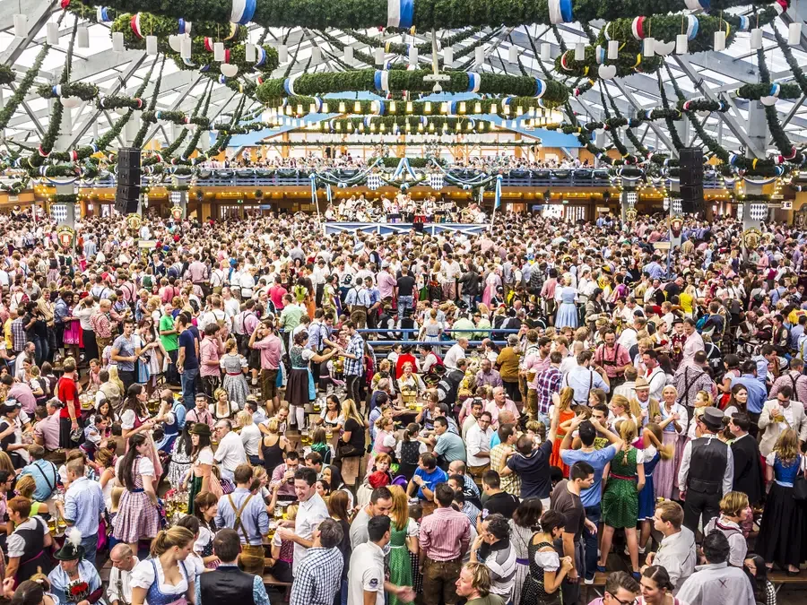 Crowd of people attending Oktoberfest in a large tent decorated with green and blue garlands and hanging ornaments.