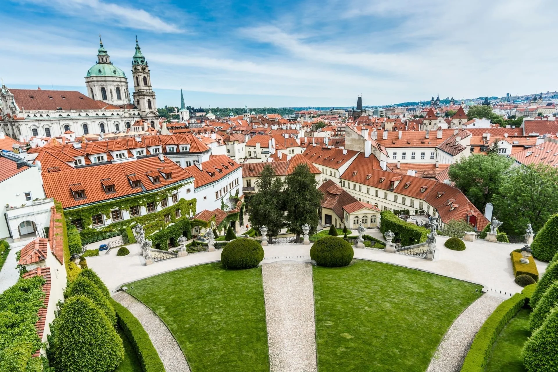 Aerial view of a historic European city with red-tiled roofs, church domes, and a lush green garden in the foreground.