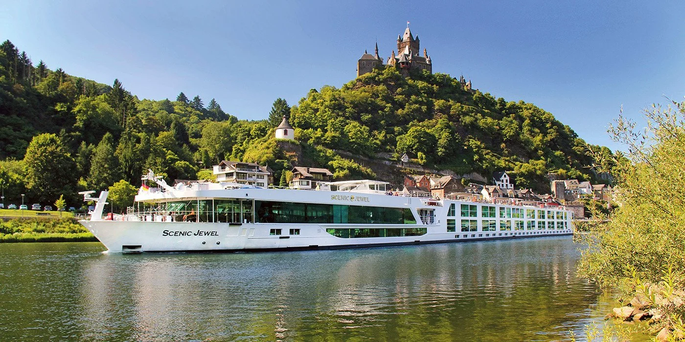 A large white cruise ship named Scenic Jewel sailing along a river with lush green hills and a castle on top in the background.