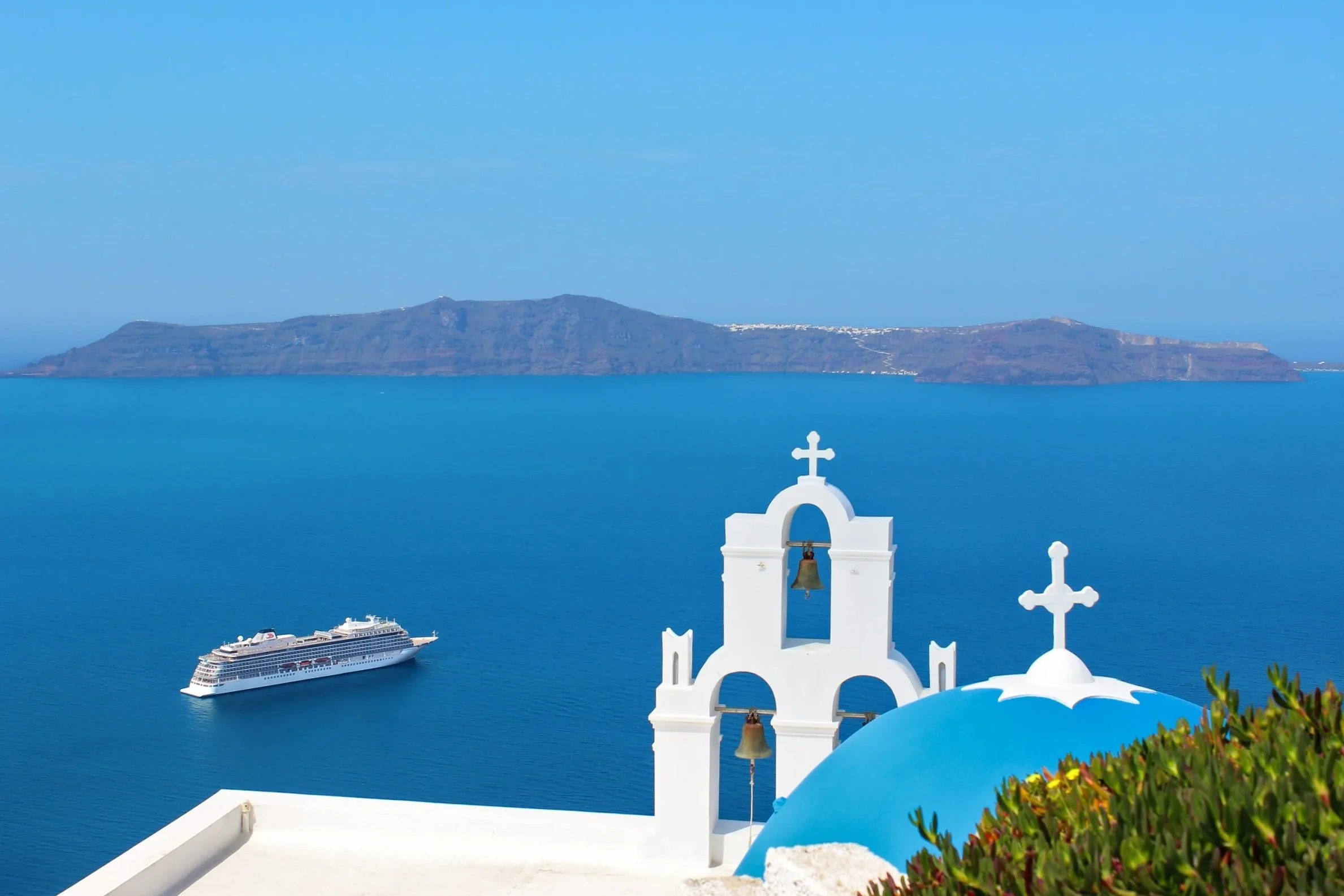 A white church bell tower with crosses, overlooking the blue waters of the Aegean Sea with a cruise ship sailing near the island of Santorini in Greece, in the background.